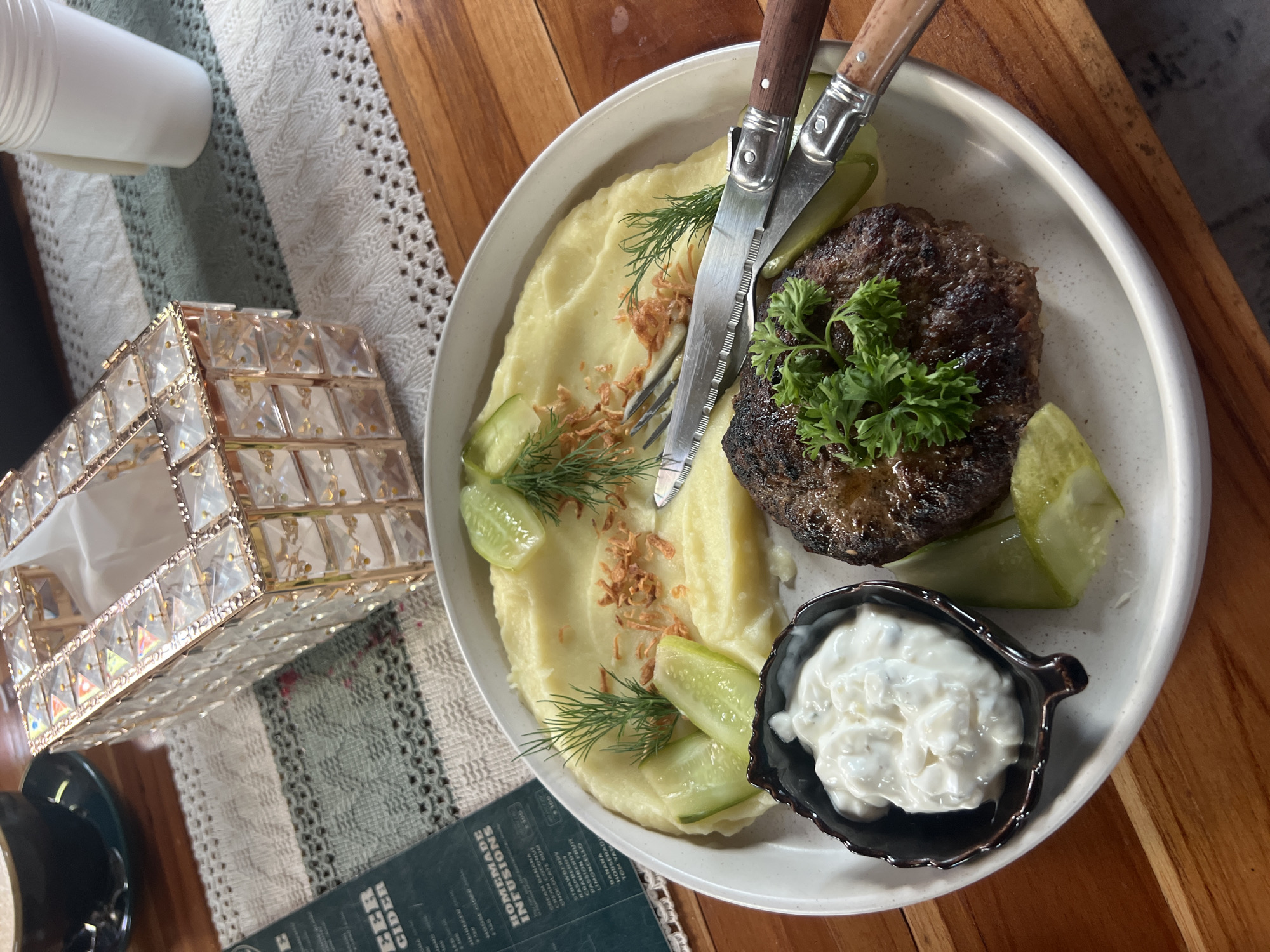 A plate of food is shown on a wooden table. The plate contains a hamburger patty, mashed potatoes, cucumber slices, dill, and a small dish of white sauce. A knife and fork are placed on the plate. To the left of the plate is a menu. In the background, a tissue box, a cup, and a napkin are visible. The overall setting suggests a meal at a restaurant or cafe. The presence of a menu with Thai script and the general ambiance are consistent with a location in Ko Samui District, Thailand. There are no people visible in the image.