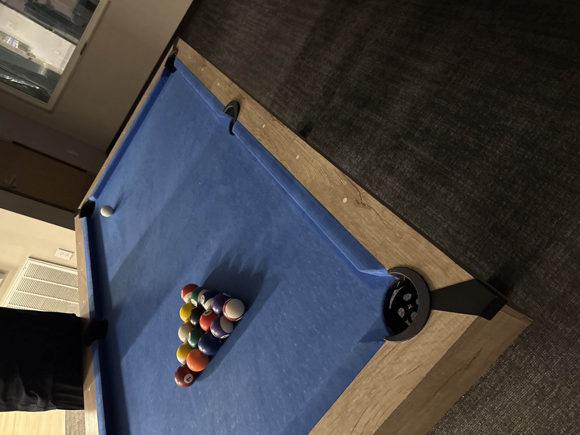 An overhead, angled shot captures a pool table set up indoors, likely in a recreation area or common room in Myrtle Beach, United States of America. The prominent subject is a modern pool table featuring a vibrant blue felt playing surface. The table is framed by light, natural wood-look side rails and has dark, possibly black, pockets and supports.
On the blue felt, a full set of billiard balls is tightly racked in a triangular formation, ready for the break shot of a game like 8-ball or 9-ball. The balls display a mix of solid colors and striped patterns, characteristic of a standard pool set. A single white cue ball is positioned at the opposite end of the table, near the head rail and a corner pocket.
The surrounding floor is covered in a dark gray or black textured carpet or rug, providing a stark contrast to the light wood and blue felt of the table. In the background, reflections are visible in what appears to be a window, suggesting artificial indoor lighting or an outdoor scene at dusk or night. A white air vent is also partially visible on a light-colored wall, indicating an air-conditioned or heated indoor environment. Part of a dark, possibly black, piece of furniture or structure is glimpsed on the left side of the frame. No people are visible, and there is no discernible text in the image. The scene suggests a paused or ready state for a game of pool.