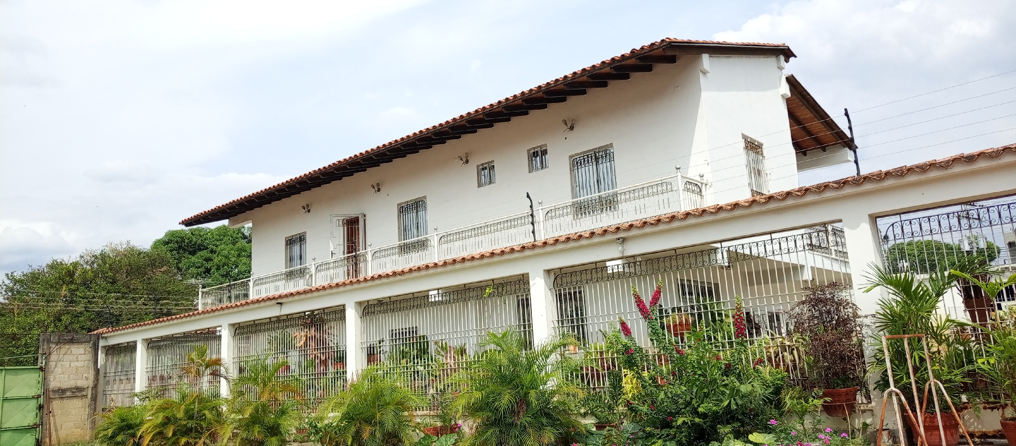 The image shows a two-story white building with a red tile roof. The upper story has a balcony with a white railing and barred windows. An open door leads to the balcony. The ground floor has a covered porch supported by white pillars. The porch is enclosed with white railings. A variety of plants and flowers are in front of the porch. A green gate is visible on the left side of the image. The building is located in Tinaquillo, Venezuela. The sky is overcast with white clouds.
