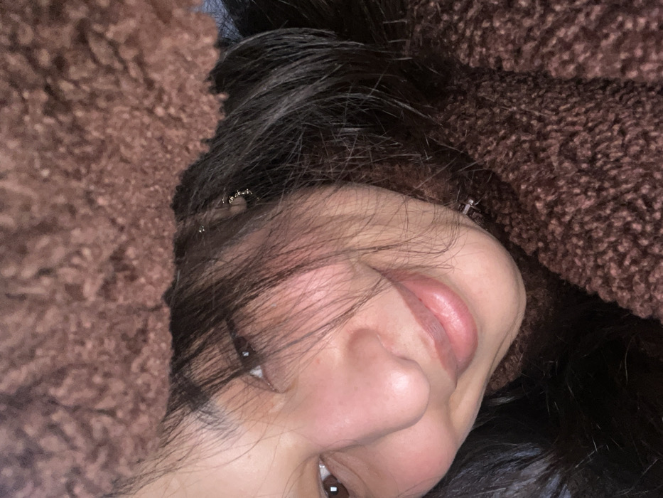 A young woman with dark hair is positioned at an angle, her face turned towards the camera. Her lips are slightly parted, and one eye is visible, reflecting light. The skin has a smooth texture. Around her face are soft, brown, textured materials, possibly clothing or blankets, that frame her. The overall impression is cozy and intimate.