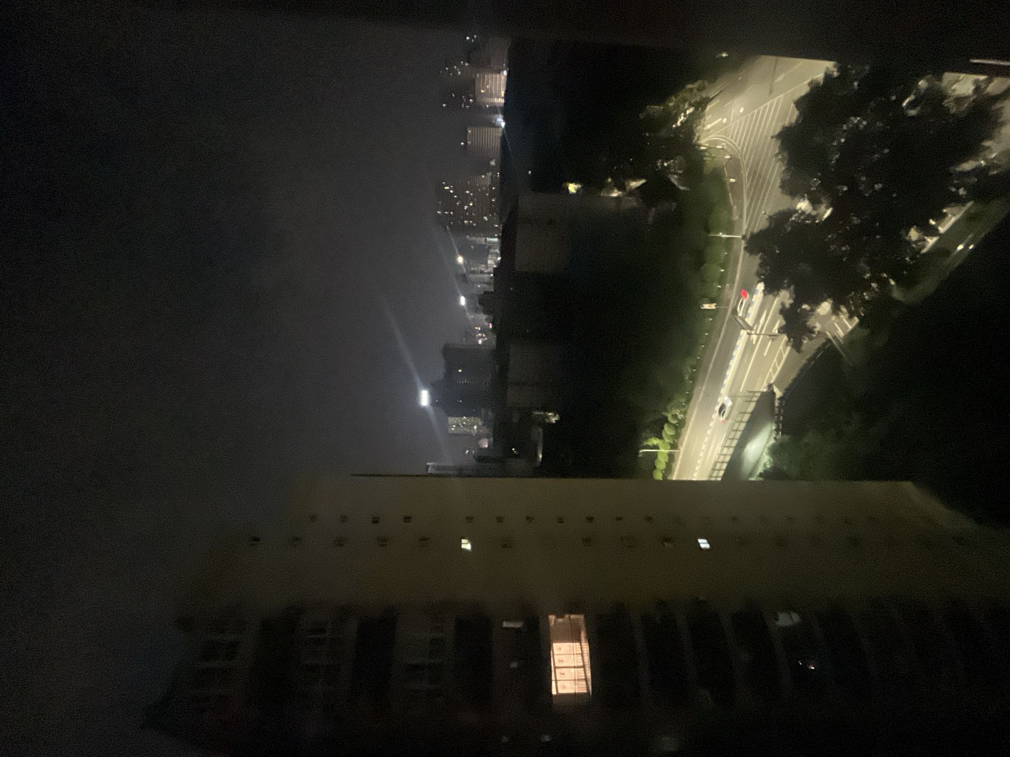 The image presents a nighttime urban scene, observed from an elevated perspective, likely within Jiulongpo District, China. A prominent, light-colored multi-story building occupies the left foreground, displaying several illuminated windows; one window on a lower floor exhibits a bright orange interior light, revealing window treatments. To the right of this building and below, a multi-lane road or highway is visible, illuminated by streetlights, with a bridge section extending across the lower part of the scene. Several vehicles are present on the road, including a white car and another vehicle displaying red taillights. Beyond the road, dense vegetation, consisting of trees and bushes, is partially illuminated by streetlights. In the midground and background, a sprawling cityscape is evident, characterized by numerous high-rise buildings with many illuminated windows, contributing to a diffuse urban glow against an overcast or hazy night sky. No individuals are discernible, and no specific actions or interactions beyond the movement of vehicles are apparent.