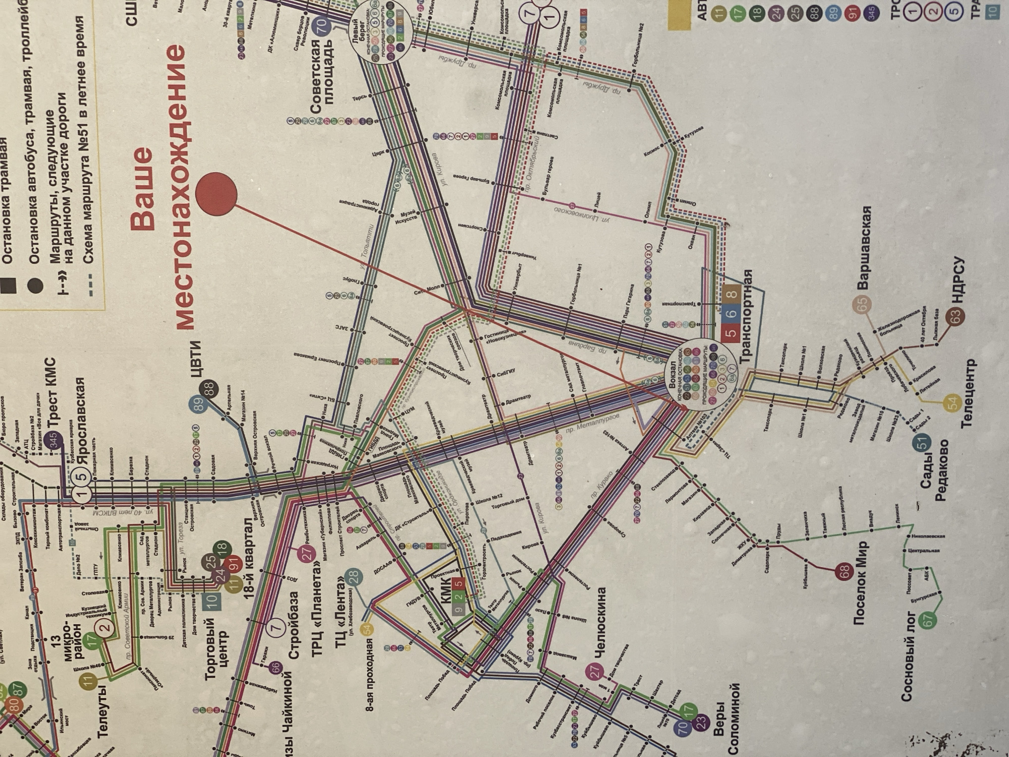 This image is a section of a public transportation map of Novokuznetsk, Russia. It details bus, tram, and trolleybus routes, with color-coded lines and numbered stops. Key locations marked include "Торговый центр" (Shopping Center), "Строибаза" (Construction Base), "Солнечная площадь" (Sunny Square), "Киселёвская" (Kiselevskaya), and "Верх-Телеутская" (Verkh-Teleutskaya). The map also features a section labeled "Ваше местонахождение" (Your Location) with a red marker, indicating the viewer's approximate position on the map. The map is in Russian, with some Cyrillic and Latin script elements. The overall condition of the map appears to be somewhat worn.