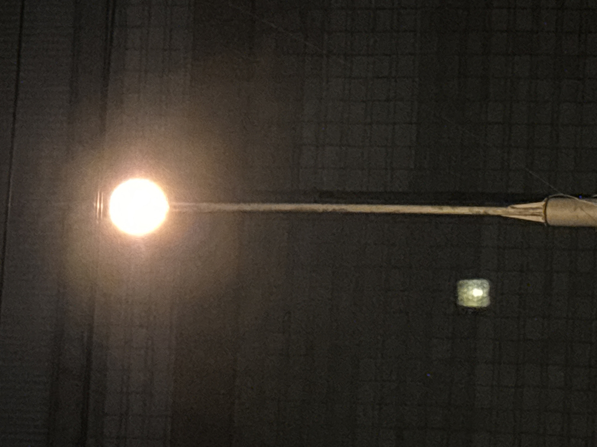 The image displays a street lamp at night, with a bright, circular light source at the top of a tall pole. The background appears to be a dark, textured wall, possibly made of brick or concrete, with a grid-like pattern. To the left of the main street lamp, a smaller, square light fixture is visible, emitting a dimmer, diffused light. The scene is devoid of any people or visible activity, suggesting a quiet, possibly urban or industrial setting. The image does not provide any specific details that would allow for the precise location within the city of Novosibirsk, Russia.