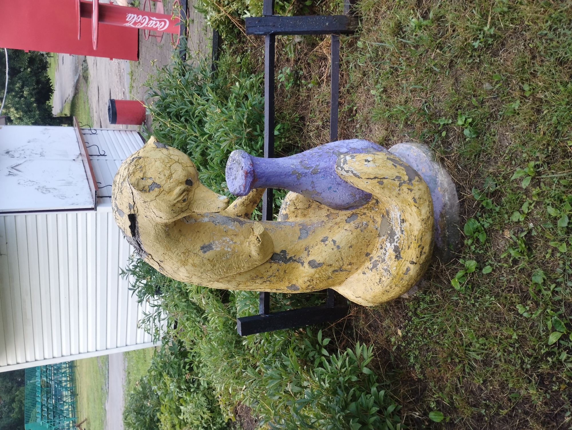 The image shows a weathered, yellow concrete sculpture of a curled-up animal, possibly a cat or a fox, with a cracked and peeling surface. The sculpture is positioned outdoors, surrounded by green foliage and grass. A purple, vase-like object is integrated into the sculpture's design, with the animal's head resting near its opening. A black metal fence with horizontal bars runs behind the sculpture. In the background, to the left, is a white building with horizontal siding, and further left, a green tiered structure resembling bleachers. To the right of the sculpture, a red Coca-Cola themed bar and seating area is visible, along with a red trash can. The scene appears to be in a public park or recreational area, possibly in the city of Sumy, Ukraine, given the context of prior images. No people are present in the image.