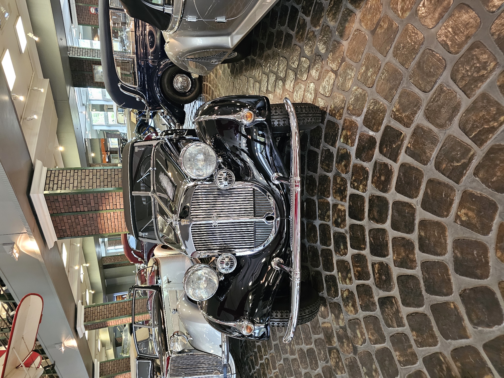 The image shows the front view of several vintage cars, with a black car prominently in the foreground, in the city of Архангельское, Russia. The cars are displayed indoors, likely in a museum or showroom, indicated by the brickwork design on pillars and ceiling structures, along with display lighting. The ground is paved with cobblestones. Other cars are partially visible behind the black one.