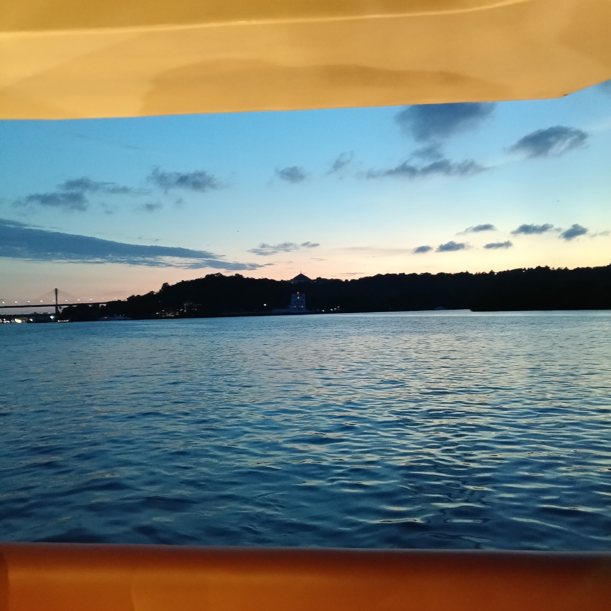 The image shows a body of water with land visible in the distance, taken from what appears to be a boat. The sky is a gradient of blue and orange, indicating either sunrise or sunset. The water has small ripples. A bridge is visible on the left side of the image. A distinct building or tower is situated in the distance, potentially on a hill or elevated land. The bottom and top parts of the image feature blurred, yellow-toned elements, likely part of the boat's structure. The scene depicts the Mandovi River in Britona, Goa, India.