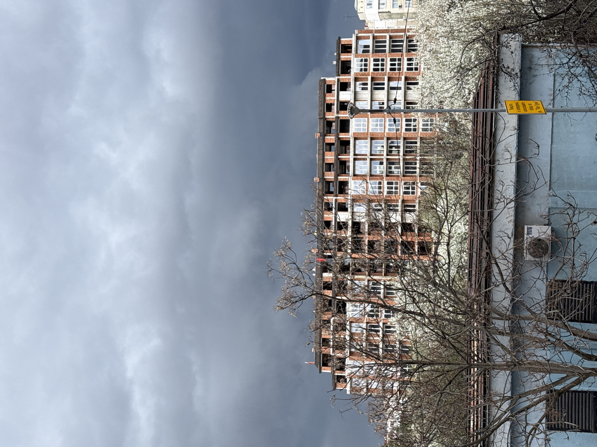 The image shows an unfinished building in Sofia, Bulgaria. The building has a brick exterior with multiple window openings. There are trees in the foreground and a gray, cloudy sky above. A light blue building is located to the right of the unfinished structure. A yellow road sign is also visible.