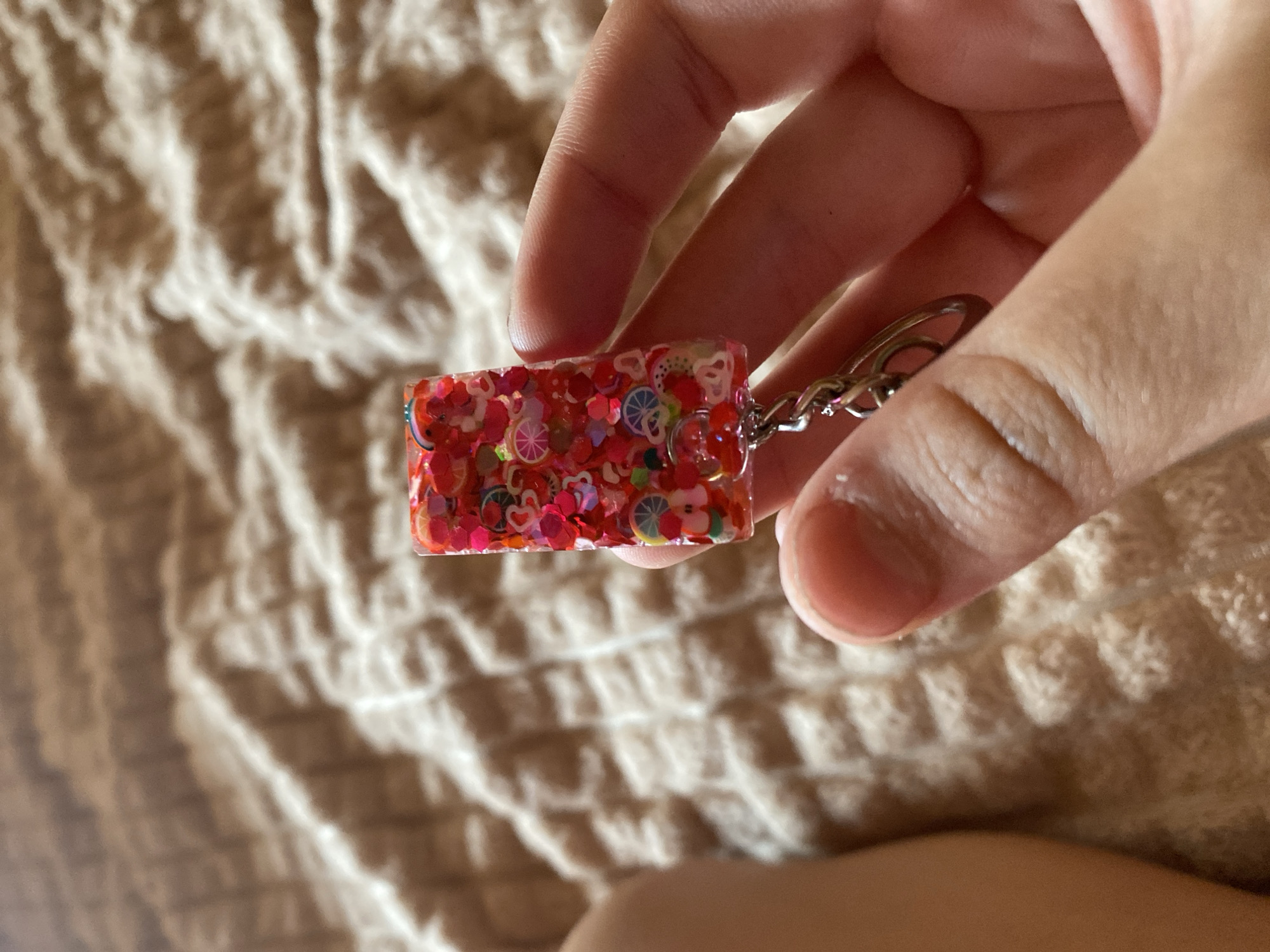 A person is holding a rectangular keychain made of clear resin. The resin is filled with small, colorful charms and glitter in shades of red, pink, and orange. Some of the charms appear to be fruit slices and hearts. The keychain is attached to a silver metal chain and a keyring. The background is a textured brown fabric, possibly a blanket or bedding. The image does not provide any information about the location in Krivodanovka, Russia.