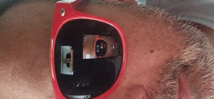 A vertical, close-up photograph depicts the lower facial area of an unidentified individual, encompassing the nose, cheeks, mouth, and chin. The subject exhibits fair skin with a distinct mole on the right cheek (viewer's left) and a short, salt-and-pepper beard. They are wearing thick, red-framed sunglasses, characteristic of a Wayfarer style.
The left lens of the sunglasses (viewer's left, subject's right eye) reflects a screen, on which a male figure in a suit and tie is visible, positioned behind what appears to be a desk or podium. A chyron or banner with text is present at the bottom of this reflected screen, partially legible as "CONSEILLE "NON NE PAS COMPLIQUER"".
The right lens of the sunglasses (viewer's right, subject's left eye) displays a multi-layered reflection. The most prominent reflection is that of the photographer holding a smartphone, which is capturing the image. Within this reflection, the red frames and lenses of the subject's sunglasses are visible again, creating a recursive visual effect. A small, circular white sticker is also discernible in the background of this reflection.
The setting is inferred to be indoors, likely within the city of Esch-sur-Alzette, Luxembourg, given the French language displayed on the reflected screen, which aligns with the linguistic context of the region. The image captures a self-portrait action while the subject is in proximity to a display screen.