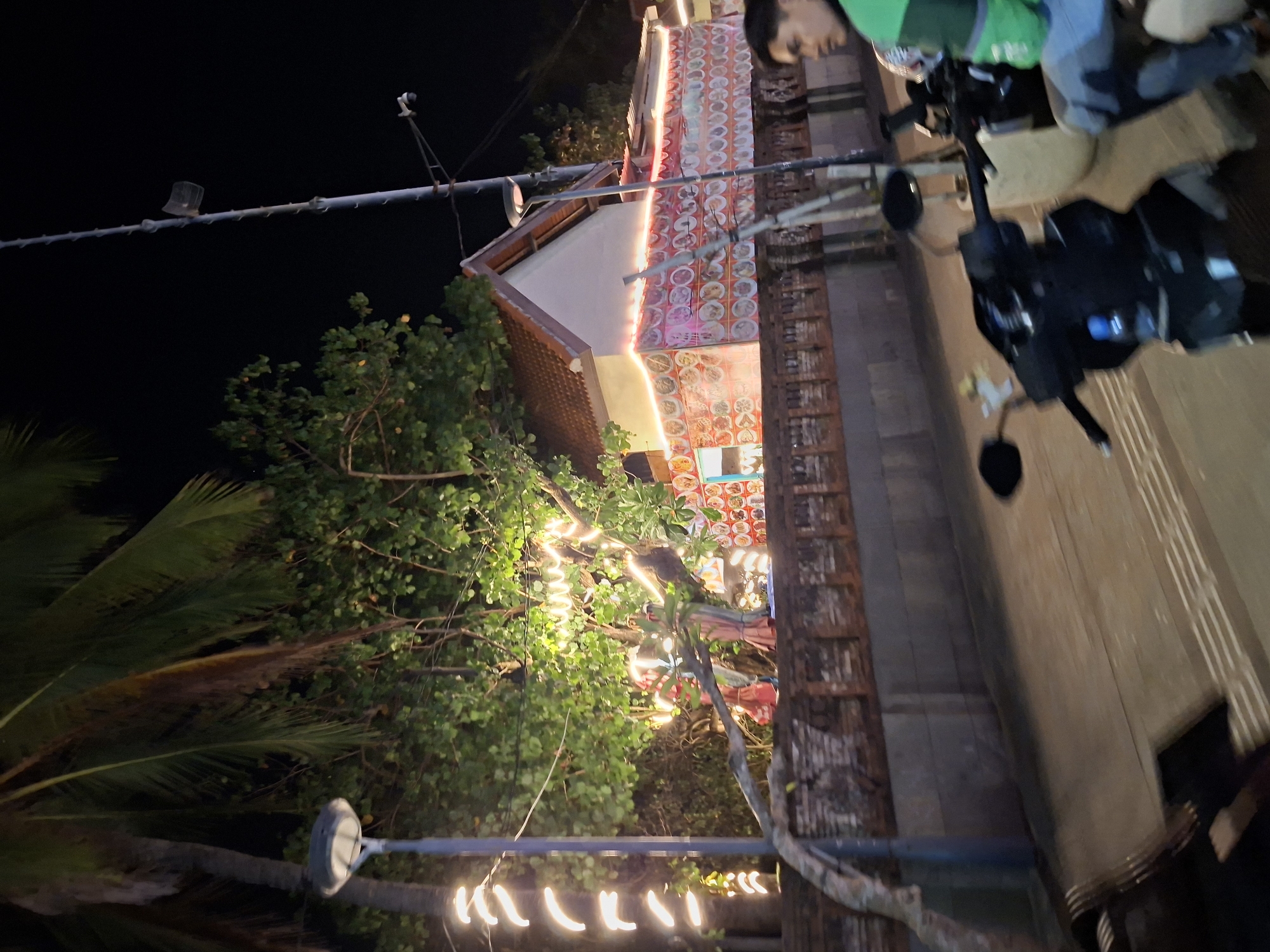 This is a nighttime street-level, slightly blurred, vertical photograph taken in Kuta, Indonesia. In the foreground, a dark-colored scooter is parked, with its handlebars and mirrors visible. A person wearing a green jacket and blue jeans is seated on a scooter to the right, looking to the left of the frame. Behind the scooters, a low brick wall runs horizontally across the middle ground. Beyond the wall, a building with a tiled roof and white walls is illuminated. The building's facade is decorated with numerous circular images, possibly of food items, and is outlined with white string lights. A large tree with green leaves, also adorned with yellow string lights, stands to the left of the building. A tall lamppost is positioned to the left of the tree, with another lamppost further back to the right. The overall scene suggests a bustling area with some nighttime commercial activity.