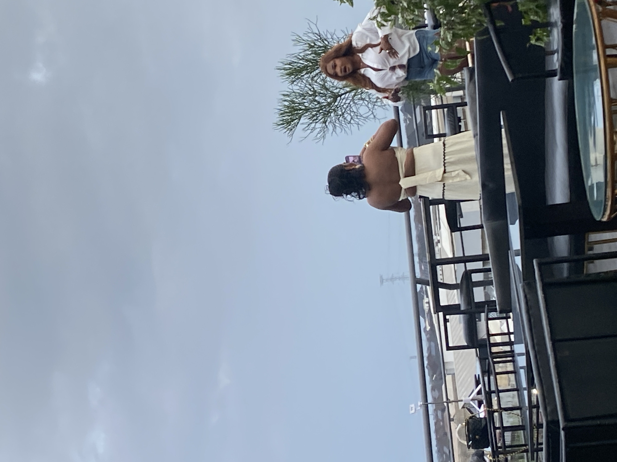Two women are on an outdoor balcony in an urban setting under a cloudy sky. The woman on the left, seen from behind, wears a strapless off-white dress with a tie detail at the back and is holding up a smartphone. The woman on the right, facing forward with her mouth open, wears a white unbuttoned shirt over a white top, blue denim shorts, and has long, reddish-brown hair. She is gesturing with her hands. In the foreground, there are dark-colored tables and seating arrangements, suggesting an outdoor dining or lounge area. In the background, there are buildings, industrial structures, and a power line tower.