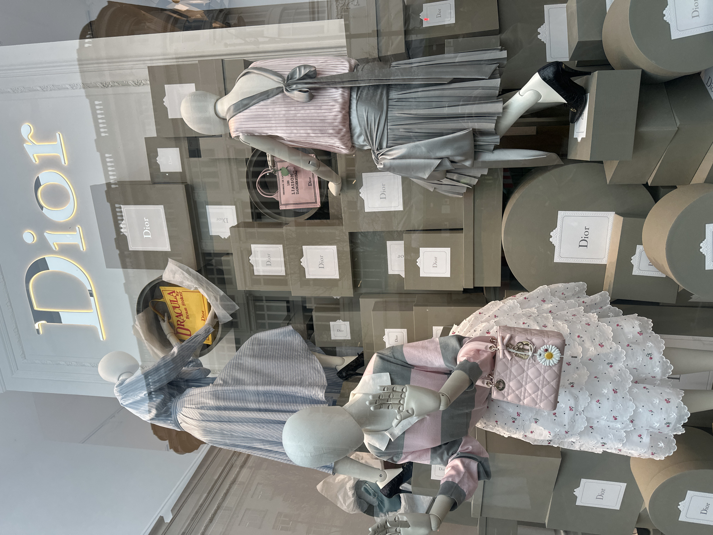 This is a street-level window display of a Dior retail store in London, United Kingdom. The scene features three white, featureless mannequins dynamically arranged among a series of stacked grey boxes and cylindrical pedestals. Most of these display elements are adorned with white labels featuring the "Dior" logo and an ornate border. A large, illuminated, metallic "Dior" sign is positioned at the top of the display.
On the left, one mannequin wears a light blue and white striped top and a voluminous matching skirt with black stiletto heels. It is posed mid-action, appearing to step onto a grey box while reaching for a yellow tote bag printed with "DRACULA By Bram Stoker" and "Dior."
In the foreground on the left, another mannequin wears a collared, long-sleeved top with horizontal pink and grey stripes, paired with a white tiered skirt embroidered with small red flowers. It carries a small pink quilted handbag accented with a white daisy charm. Its articulated hands are raised with palms open.
On the right, a third mannequin is dressed in a sleeveless, vertically-pleated pink top with a grey sash tied in a large bow at the back, a grey pleated skirt, and black stiletto heels. Near this mannequin, a pink canvas tote bag, labeled "LES LIAISONS DANGEREUSE Dior," is displayed on a grey box. Another black crocodile-embossed stiletto heel with a gold buckle detail rests on a separate grey box in the lower right foreground.
Reflections in the display glass reveal architectural details of buildings across the street, consistent with classical London street architecture, including ornate window frames and building structures.