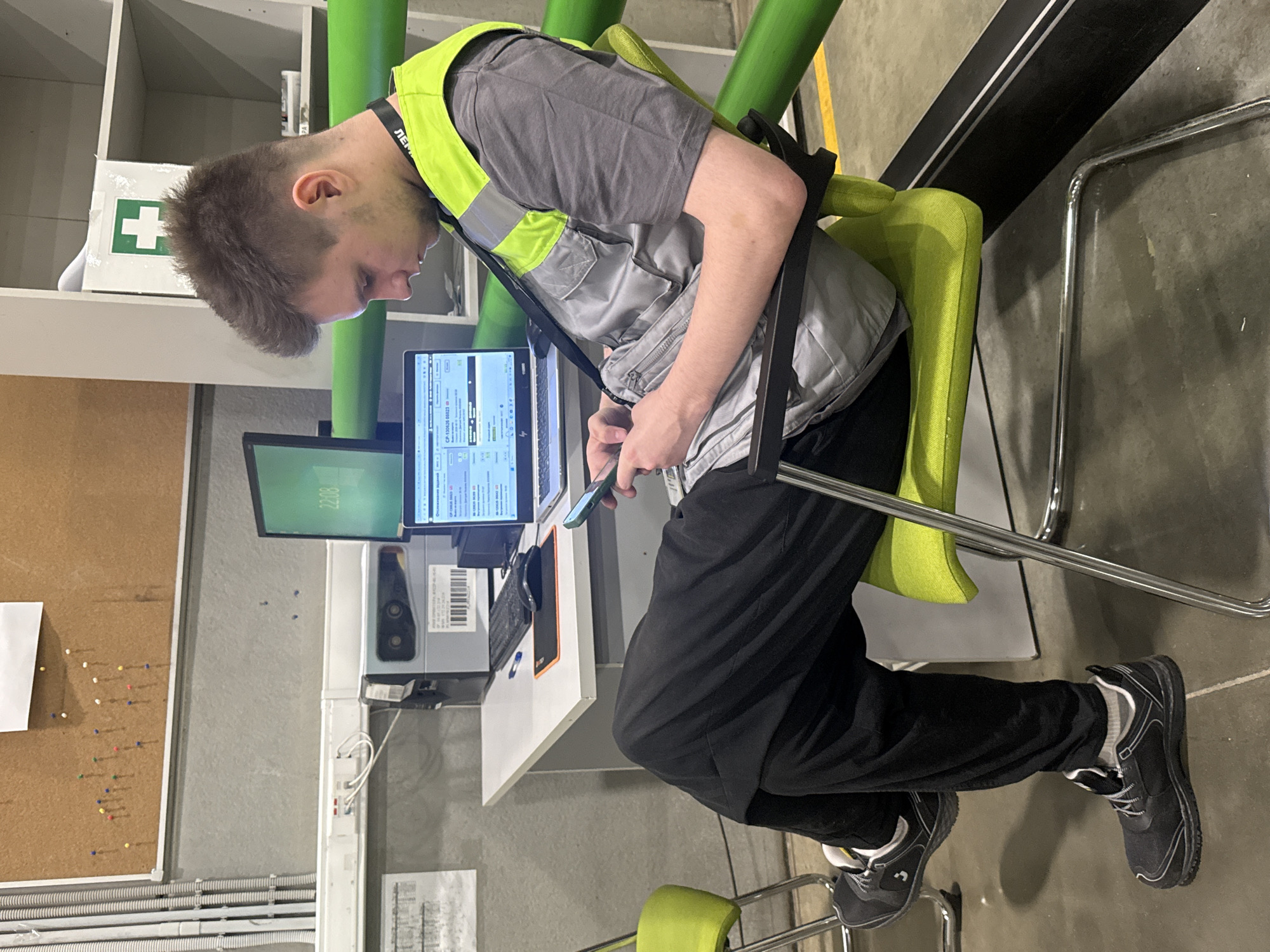 A young man with short brown hair, wearing a grey t-shirt, a grey work vest with reflective yellow trim, black pants, and black work shoes with grey accents, is seated in a lime green chair at a white desk. He is looking down at a smartphone in his hands. On the desk is an open laptop displaying a program with Russian text, a keyboard, a mouse, and a pen. Behind the desk, a computer monitor displays the time "22:08". To the left of the desk, a bulletin board is pinned with numerous colored pushpins. In the background, a white shelf unit with a green cross medical symbol on a white box is visible. The environment appears to be an industrial or workshop setting, with concrete floors, grey walls, and large green pipes running horizontally. The location is identified as Lupanovo, Russia.