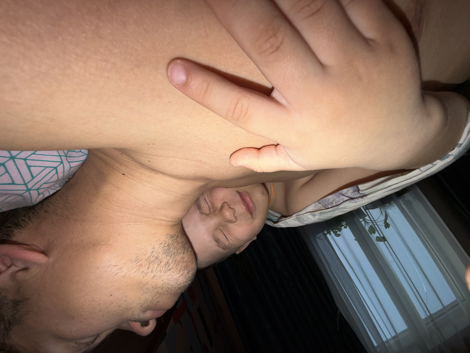 A partially lit, close-up, eye-level shot depicts two individuals in a domestic indoor setting.
A man with dark hair and facial stubble, lying on his side, rests his head on a light-skinned child's chest. The child lies on his back with eyes closed and a hand resting on his torso. Light colored bed sheets and pillows are partially visible.
The background contains dark walls and a window covered with sheer white curtains. A potted plant sits on the windowsill.
The location of the image is unidentifiable as being in Chaikovsky, Russia.
