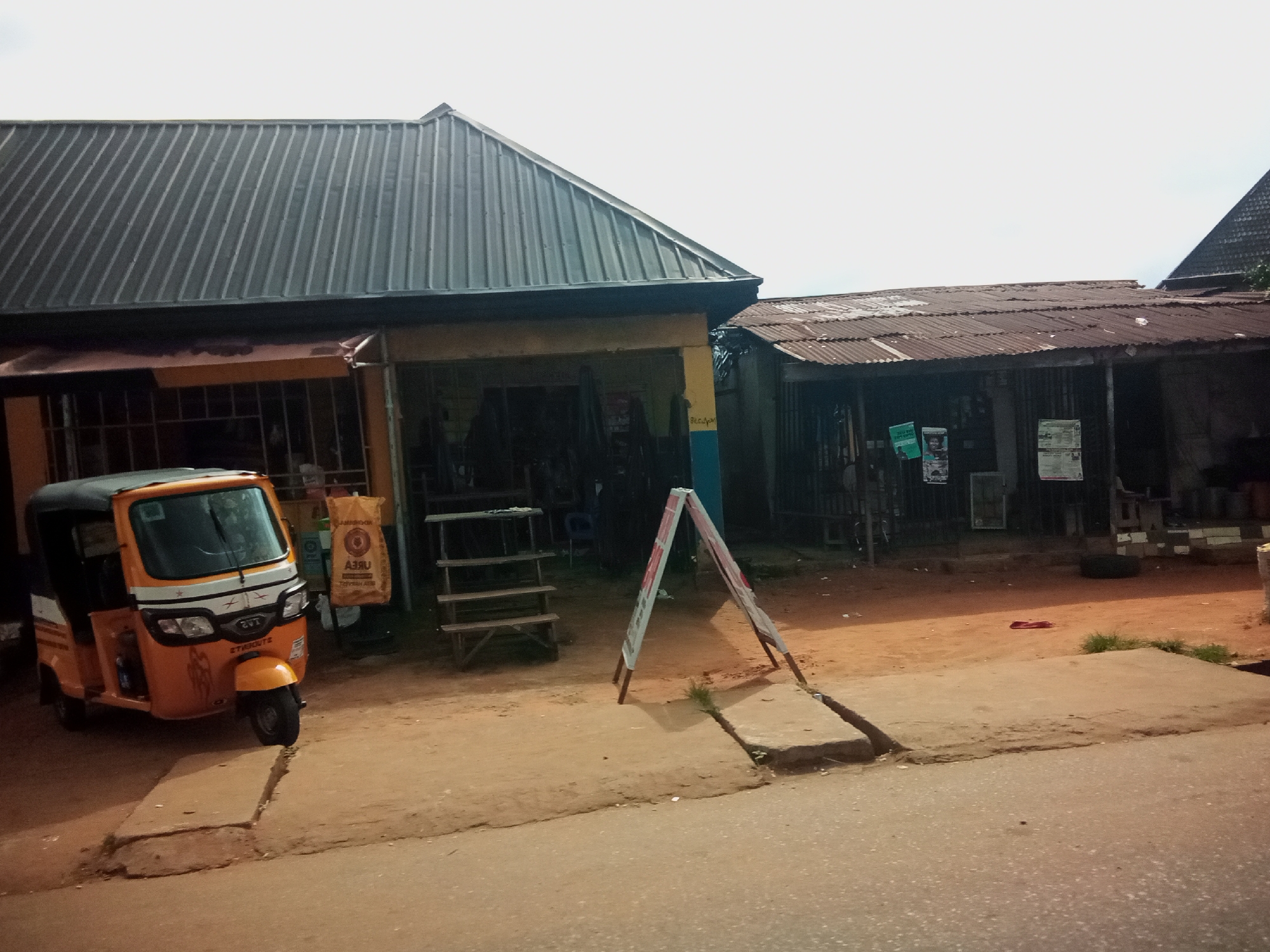 The image depicts a street scene in Nibo, Nigeria, featuring a collection of small shops and a three-wheeled vehicle. In the foreground, a bright orange auto-rickshaw, often referred to as a 'keke' in Nigeria, is parked on a dusty, unpaved surface adjacent to a paved road. The auto-rickshaw has a covered passenger section and an open driver's area. Behind the auto-rickshaw are several storefronts. The leftmost building has a corrugated metal roof and an open-fronted design with bars. To its right, another building with a similar corrugated metal roof appears to be a workshop or store, with various items visible inside, including what looks like metal equipment. Further to the right, a third building has a rusty, corrugated metal roof, suggesting an older structure. In front of these shops, a wooden display stand with several shelves is visible, and an A-frame sign with red lettering stands prominently. The ground is a mix of sand and dirt, with some patches of green grass. The sky is overcast, indicating a cloudy day. The overall impression is a typical roadside commercial area in a Nigerian town.