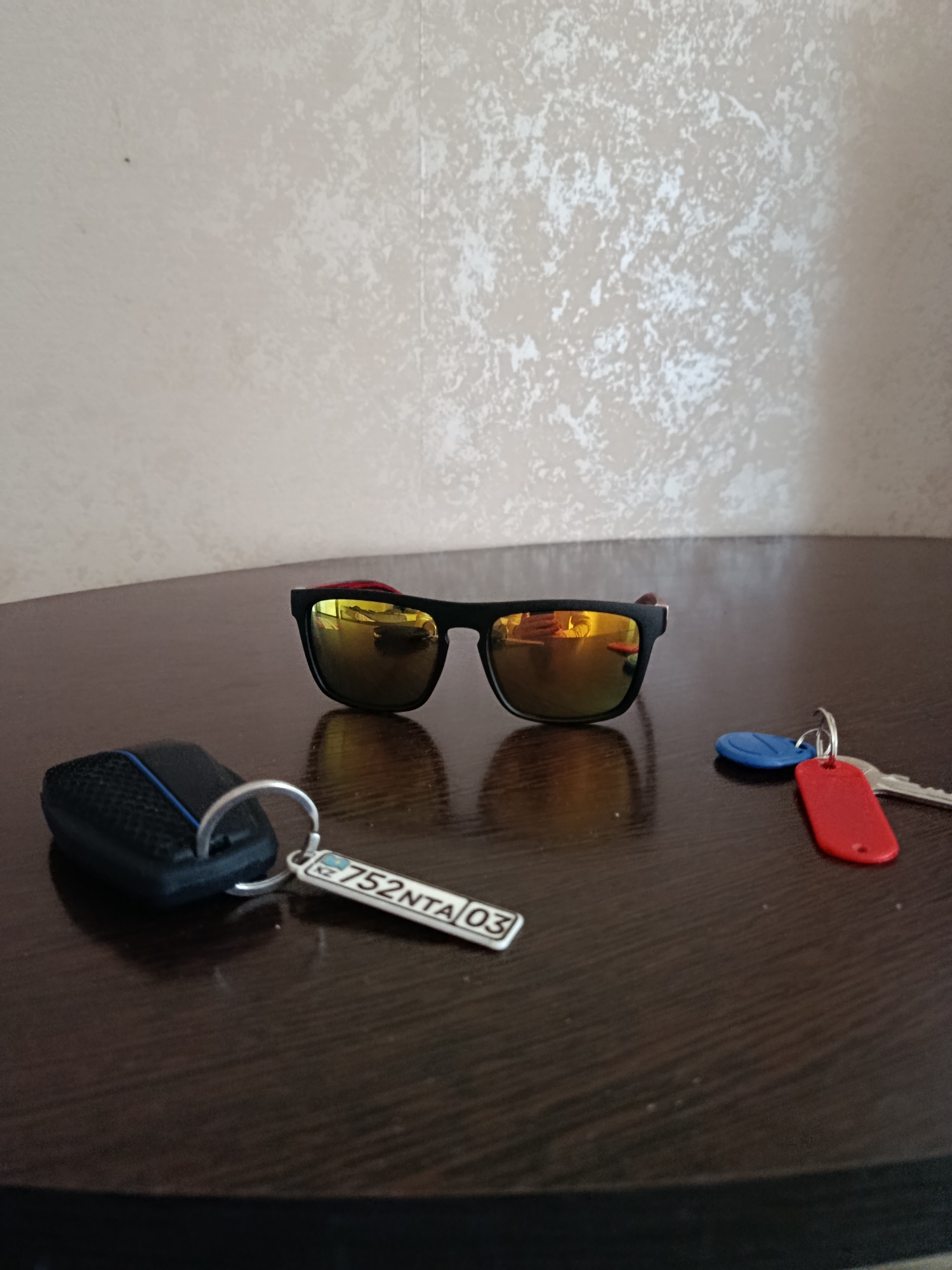 The image depicts a close-up, eye-level shot of several objects on a dark, polished wooden surface. In the foreground, a black car remote key fob with a textured surface and a blue stripe is attached to a silver keyring. Next to it, on the same keyring, is a rectangular white tag with the alphanumeric characters "752NTA 03" and a KZ symbol. To the right of the car key, a set of two keys with colored keycaps is present: one with a red keycap and another with a blue keycap. Dominating the center of the frame are a pair of black-framed sunglasses with mirrored, golden-tinted lenses, which reflect a blurry image of a person's hands and a red object. The background consists of a textured, light-colored wall. The scene appears to be indoors, likely on a table or desk. There is no discernible connection to Petropavl, Kazakhstan, beyond the presence of the KZ symbol on the key tag.