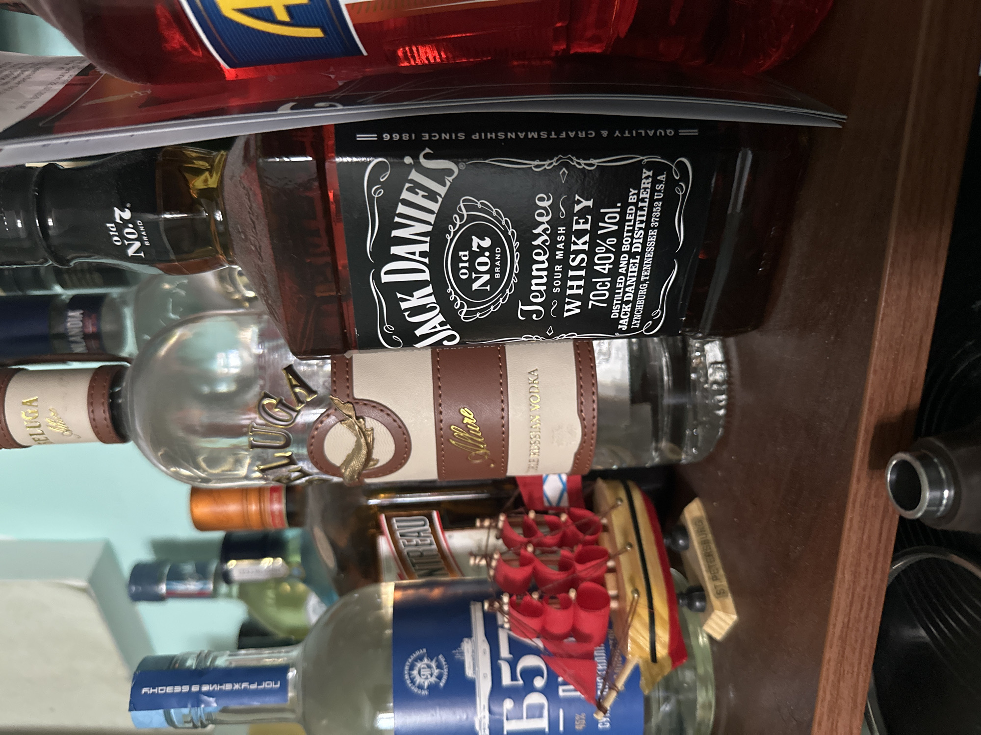 The image shows a close-up of various bottles of alcohol displayed on a wooden shelf. In the foreground, a bottle of Jack Daniel's Tennessee Whiskey is prominently featured. To the left of the Jack Daniel's bottle, a bottle of Beluga Allure Russian Vodka is visible, with a small wooden boat model placed in front of it. Further to the left, part of another clear bottle with a blue label that reads "B57" is visible. Behind these bottles, other bottles are partially obscured, including what appears to be a bottle of Cointreau and a clear bottle with a white label. A bright red bottle is visible on the far right. The shelf itself is made of wood and appears to be part of a home bar or collection. The background shows a wall with a pale blue hue. The image is taken from a low angle, focusing on the bottles. There is no information within the image to suggest a specific location within Vladivostok, Russia.