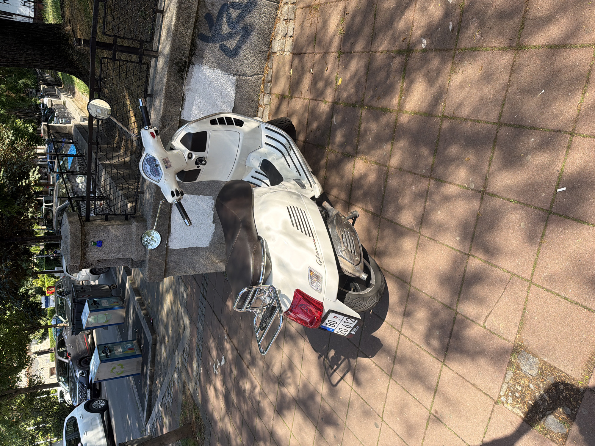 A white Vespa GTS 300 Super scooter with a brown seat and chrome rear rack is parked on a reddish-brown tiled sidewalk, partially illuminated by direct sunlight and dappled shadows from overhead trees. The scooter bears a Serbian license plate "BG 2D-612," indicating its registration in Belgrade. To the left, a street is visible with several parked vehicles, including a white five-door hatchback and a dark grey SUV, along with two blue and white recycling bins and a standard grey refuse bin near the curb. A low stone wall with a white painted section featuring black graffiti lines the right side of the sidewalk, supporting a black metal fence in the background. A small blue frog figurine is attached to a stone pillar on this wall. The sidewalk shows grass growth between the tiles and a discarded cigarette butt. No individuals are present in the scene, which depicts an urban street environment in Belgrade, Serbia.