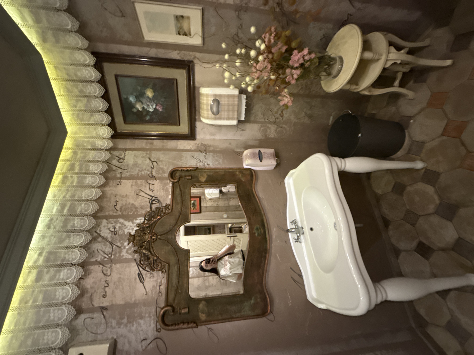 The image captures a detailed interior view of a bathroom, characterized by its vintage design elements. A white pedestal sink with chrome fixtures is prominently featured. To the left of the sink, an ornately framed mirror reflects a person holding a phone, suggesting they are taking a picture. Above the sink and mirror, there's a small framed painting with a dark wood frame, along with a second, smaller, framed print. A decorative flower arrangement sits atop a two-tiered table. The wallpaper is a muted brown, adorned with faded calligraphy and script.
The floor is tiled with a pattern of irregular hexagonal shapes in earth tones. A dark container, possibly a wastebasket, is placed near the table. Based on the information available and the vintage design aspects, the location appears to be in Sofia, Bulgaria.