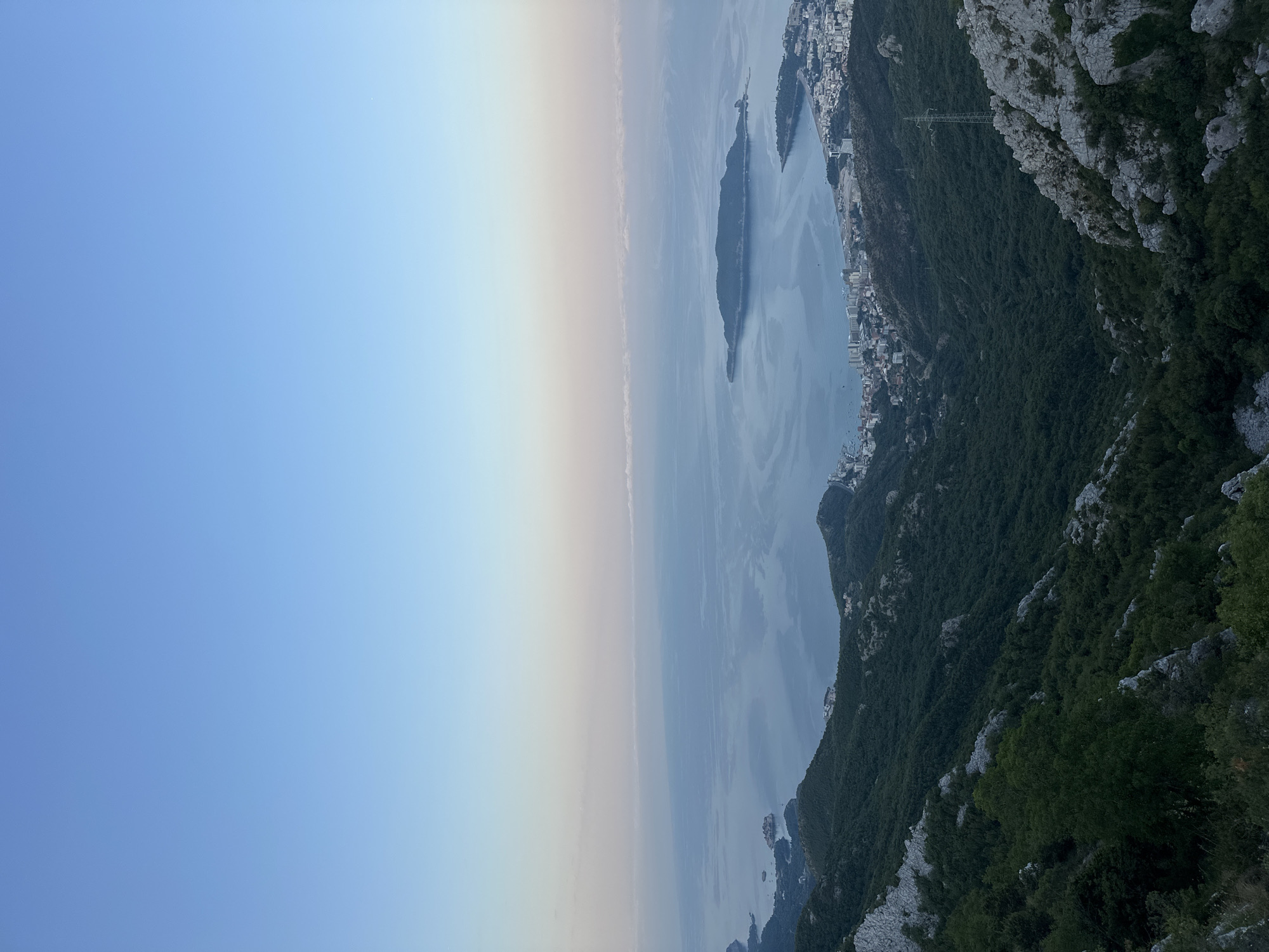 This is a wide-angle, elevated shot of the Adriatic Sea coastline near Brajici, Montenegro, taken from a mountainous vantage point. The sky is a clear, gradient blue, transitioning from a pale cerulean at the top to a soft pink and orange near the horizon, indicating either sunrise or sunset.
Below the sky, the tranquil sea stretches out, reflecting the muted colors of the sky. A long, narrow island, covered in lush green vegetation, is prominent in the mid-ground of the water. Closer to the shore, a coastal town with white buildings and a dense urban layout is visible. The coastline curves, revealing more of the town nestled between the mountains and the sea.
The foreground is dominated by steep, verdant mountains. Dense green trees and shrubs cover the slopes, interspersed with patches of gray, rocky outcrops and exposed stone. A narrow, winding road or path is barely discernible on one of the mountain slopes. The overall impression is one of a serene, natural landscape meeting a developed coastal area. There are no people or animals visible in the frame.