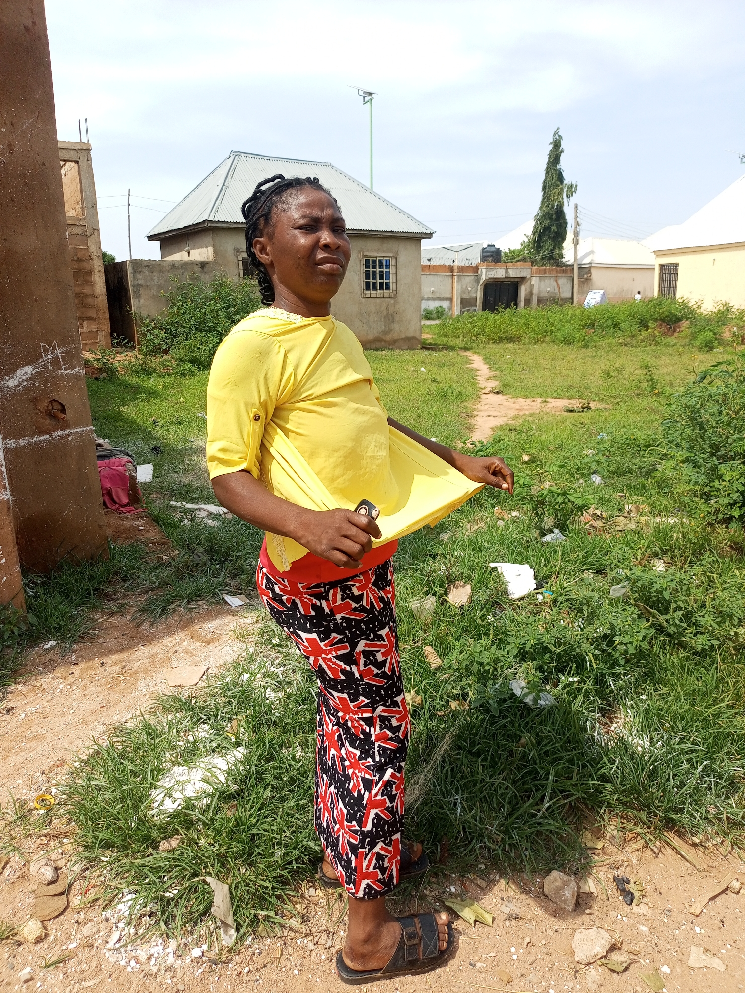 A medium shot captures a woman standing outdoors in what appears to be a residential area in Gombe, Nigeria. She is positioned facing slightly away from the camera, her head turned to her left, and her gaze directed upwards and to her left. She has dark skin and her hair is styled in braids. She is wearing a bright yellow, short-sleeved shirt, the hem of which she is holding up with her left hand to reveal a glimpse of what might be a light-colored garment underneath, and a long, patterned skirt with red, black, and white designs. Her right hand is holding a small, dark object, possibly a mobile phone.
The environment is somewhat unkempt. To the woman's left, a partially constructed building made of concrete blocks and brown plaster is visible. Beyond this, there are other buildings with corrugated metal roofs and concrete walls, indicative of typical residential structures in the area. The ground is a mix of dirt and sparse, dry grass, with some scattered debris, including white plastic bags. A narrow, sandy path winds through the grass in the background. The sky is overcast with scattered clouds. The overall impression is one of a warm, possibly dry climate, with everyday life unfolding in a semi-urban setting.
