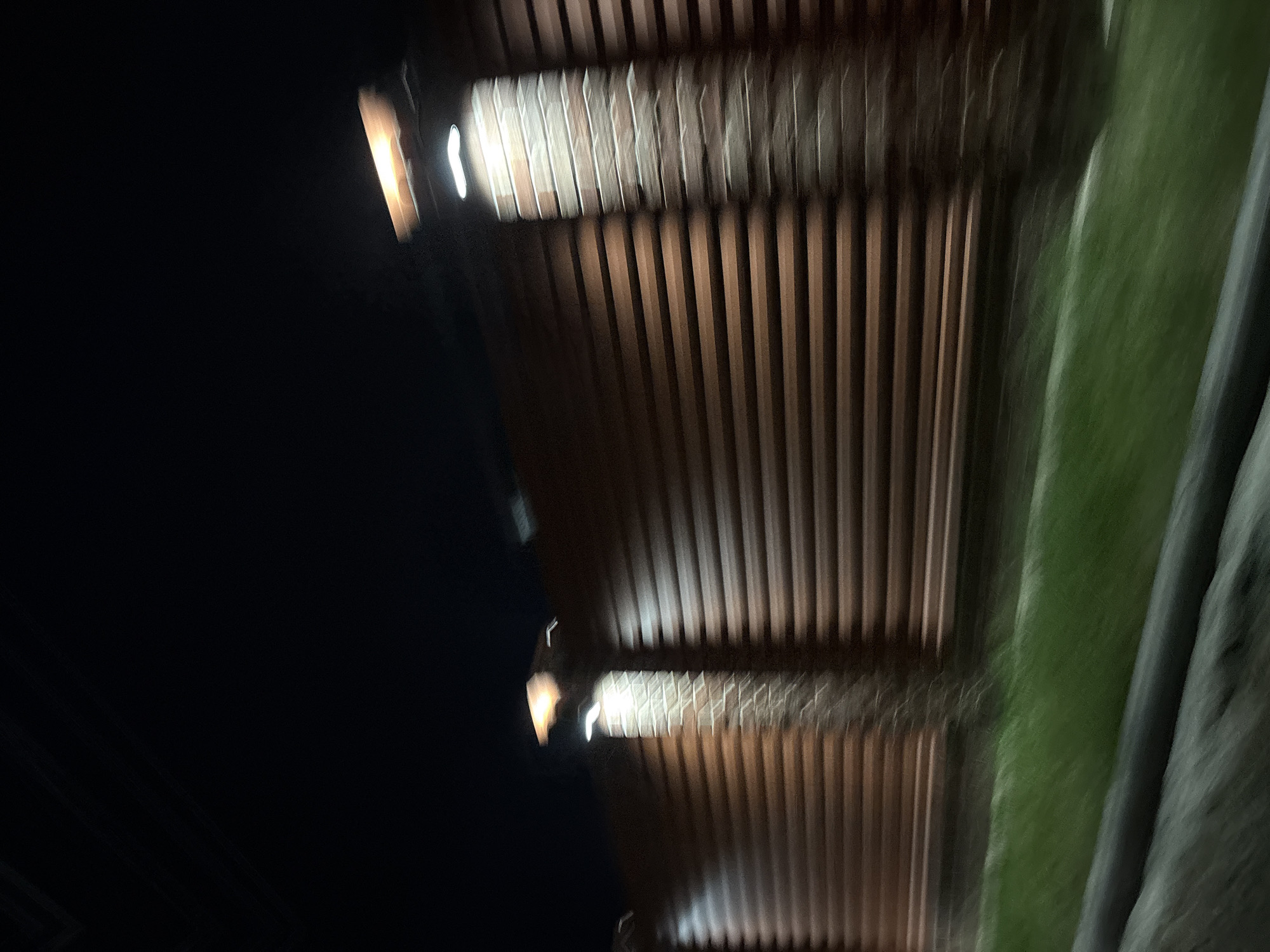 The image depicts a brick wall with horizontal siding and illuminated pillars at night. The lighting appears to be from artificial sources mounted on the pillars, casting a warm glow on the wall. A strip of green grass is visible in the foreground, suggesting an outdoor setting, possibly a road or pathway adjacent to the wall. The overall impression is one of a constructed barrier in a dimly lit environment. The image does not provide any specific visual cues to place it within the city of Agronom, Russia.