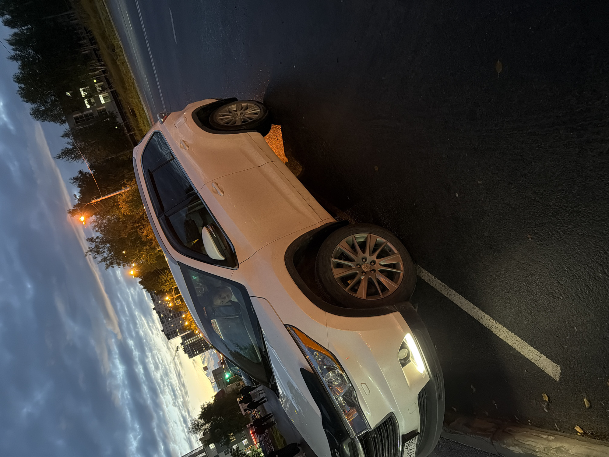 The image shows a white SUV parked on a wet asphalt road in Nizhnevartovsk, Russia, at dusk. The sky is cloudy with streaks of orange and yellow light from the setting sun. Trees and apartment buildings are visible in the background, illuminated by streetlights and interior lights. A person is visible inside the driver's seat of the SUV. The car's headlights are on. Yellow leaves are scattered on the ground near the car, suggesting it is autumn. A white parking line is visible on the road.