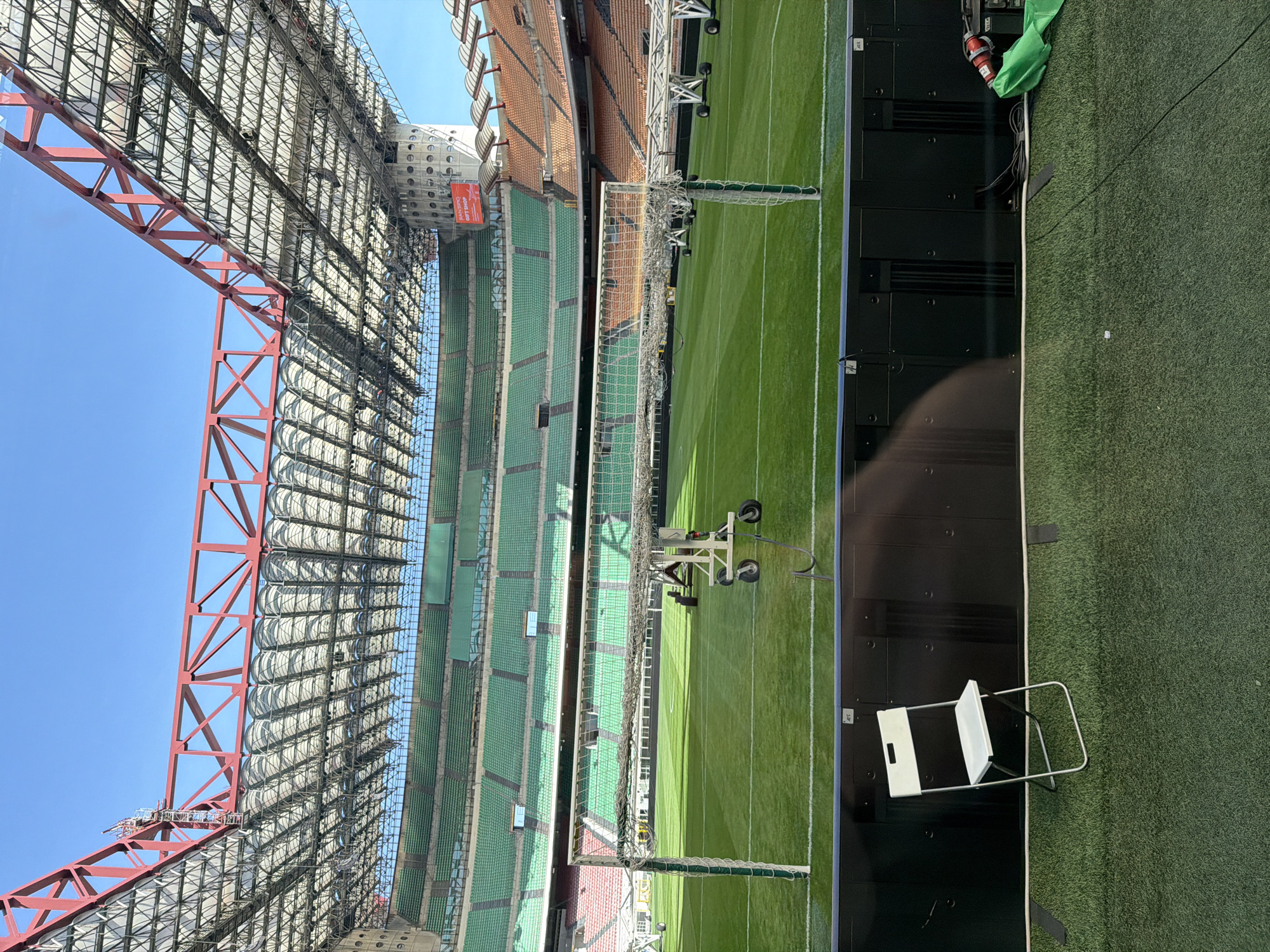 The image shows the interior of San Siro stadium in Milan, Italy. The stadium is a multi-level structure with red support beams under the roof, green and orange seats. A well-maintained green pitch occupies the center. There's camera equipment on the field. A wall and a chair are in the foreground.