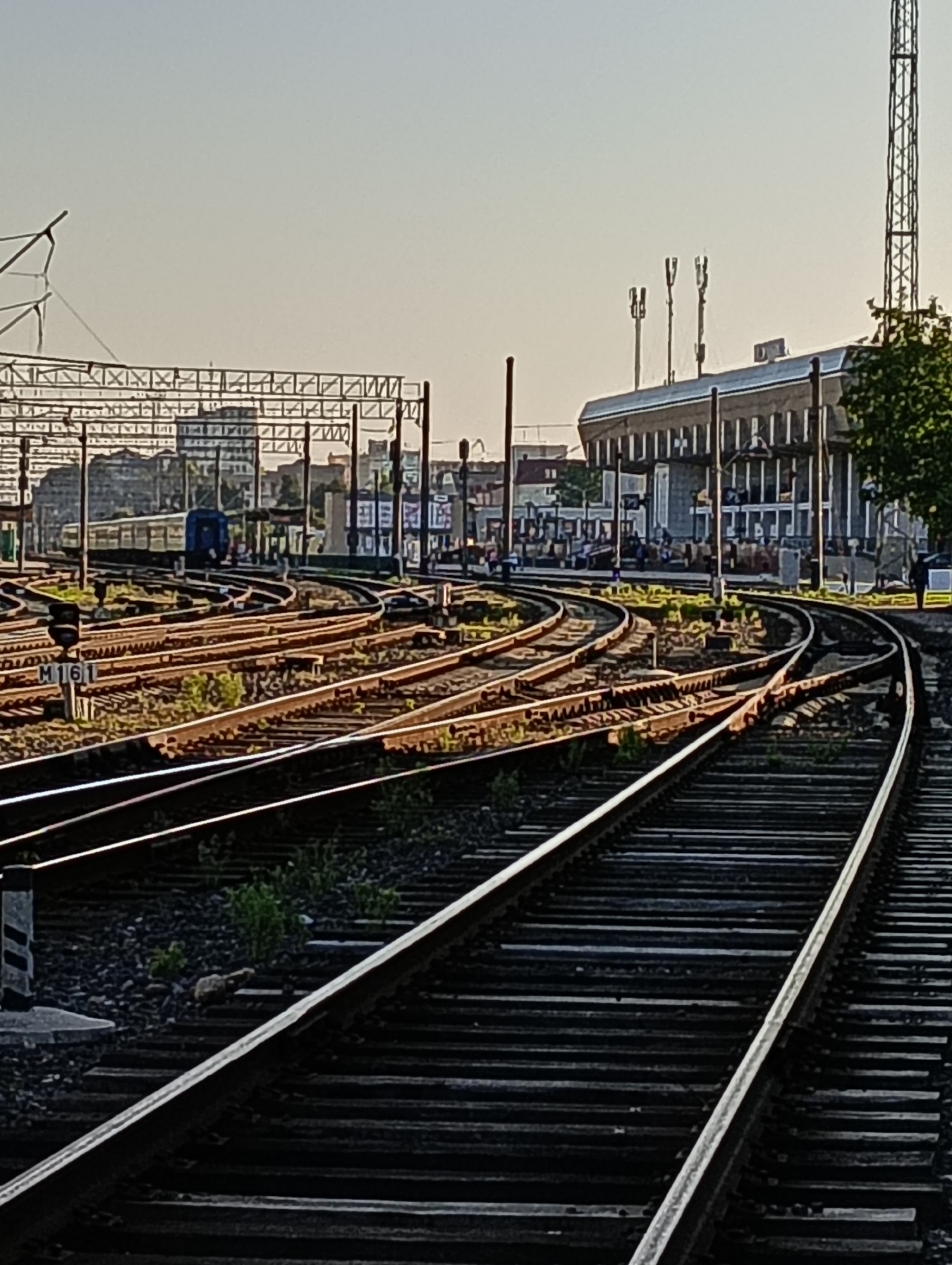 The image depicts a railway yard with multiple sets of tracks converging and diverging. In the background, a modern train station building with a glass facade is visible, alongside several cellular antennas atop the structure and a tall communication tower to its right. A train car is stopped on one of the tracks. The scene is bathed in the warm light of what appears to be either sunrise or sunset, casting long shadows. The foreground is dominated by the intricate network of railway tracks and ballast. This appears to be the railway station in Hrodna, Belarus, given the visual cues of station architecture and typical railway infrastructure found in Eastern Europe.