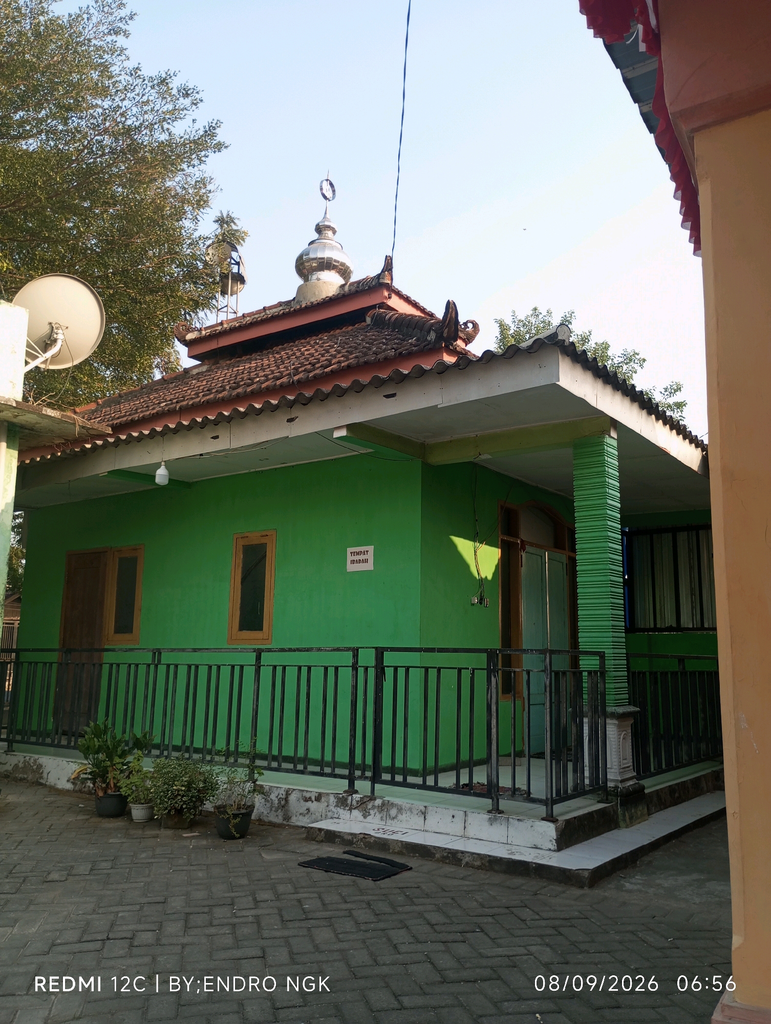 A low-angle outdoor shot captures a vibrant green building with a traditional tiled roof, likely a mosque or similar place of worship, situated in Guyangan, Indonesia. The building features dark-colored wooden doors and windows, and is partially obscured by a beige-colored structure on the right. A black metal fence runs along the front of the building, with several potted plants placed behind it. A satellite dish is visible on the left side of the building, and a single lightbulb hangs from the overhang. The sky is clear and blue, suggesting a bright, sunny day. A digital timestamp at the bottom right of the image reads "08/09/2026 06:56".