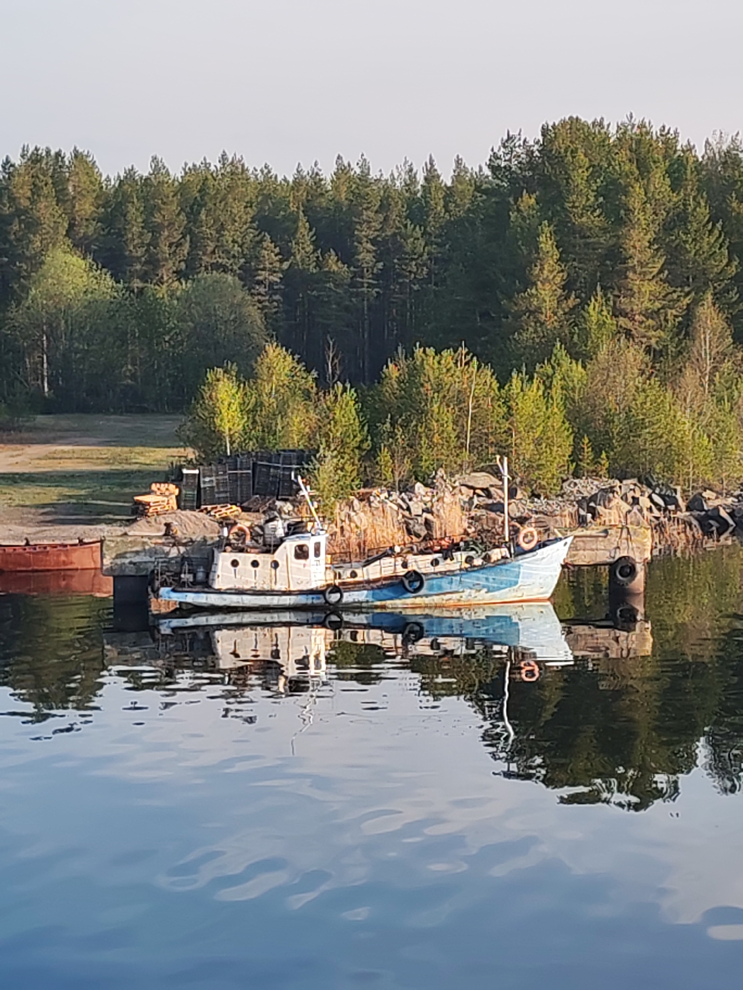 A slightly dilapidated white and blue boat is moored next to a rocky shore in Shal'skii, Russia. The boat is reflected in the calm water. The shore is littered with rocks, tires, wooden pallets, and metal crates. Behind the shore is a dense forest of green trees. A portion of a maroon-colored cylindrical tank is visible to the left of the boat.