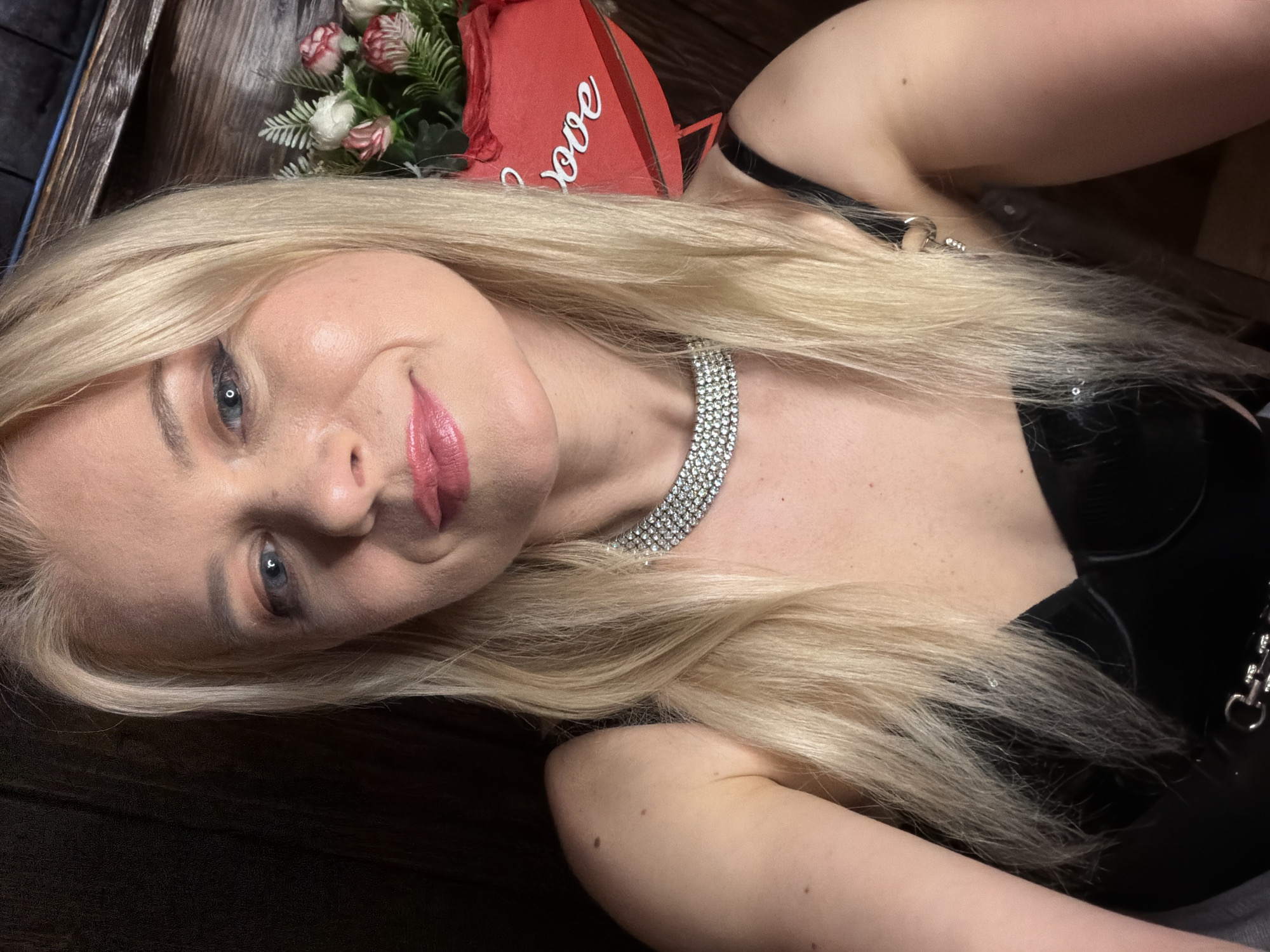A blonde woman with blue eyes and pink lipstick is taking a selfie. She is wearing a black top and a diamond necklace. Behind her, there is a heart-shaped box filled with flowers. The background appears to be a wooden wall. The image does not provide any information about the city of Sochi, Russia, or any specific location.