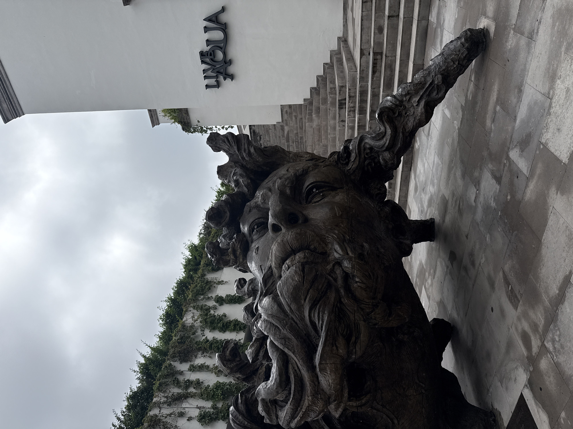 The image captures a low-angle, outdoor shot of a large, dark, sculptural head with flowing, abstract forms that suggest hair or a headdress. The sculpture is positioned on a stone-tiled ground that appears wet, indicating recent rain or a damp environment. To the right of the sculpture, a white building with a prominent black sign reading "LIVE AQUA" is visible. A wide, stepped staircase ascends the side of the building. Green ivy or vines cascade down a white wall behind the sculpture. The sky is overcast, with a pale, diffused light. The overall atmosphere is somber and artistic. This scene is located at Live Aqua Urban Resort San Miguel de Allende, Mexico.