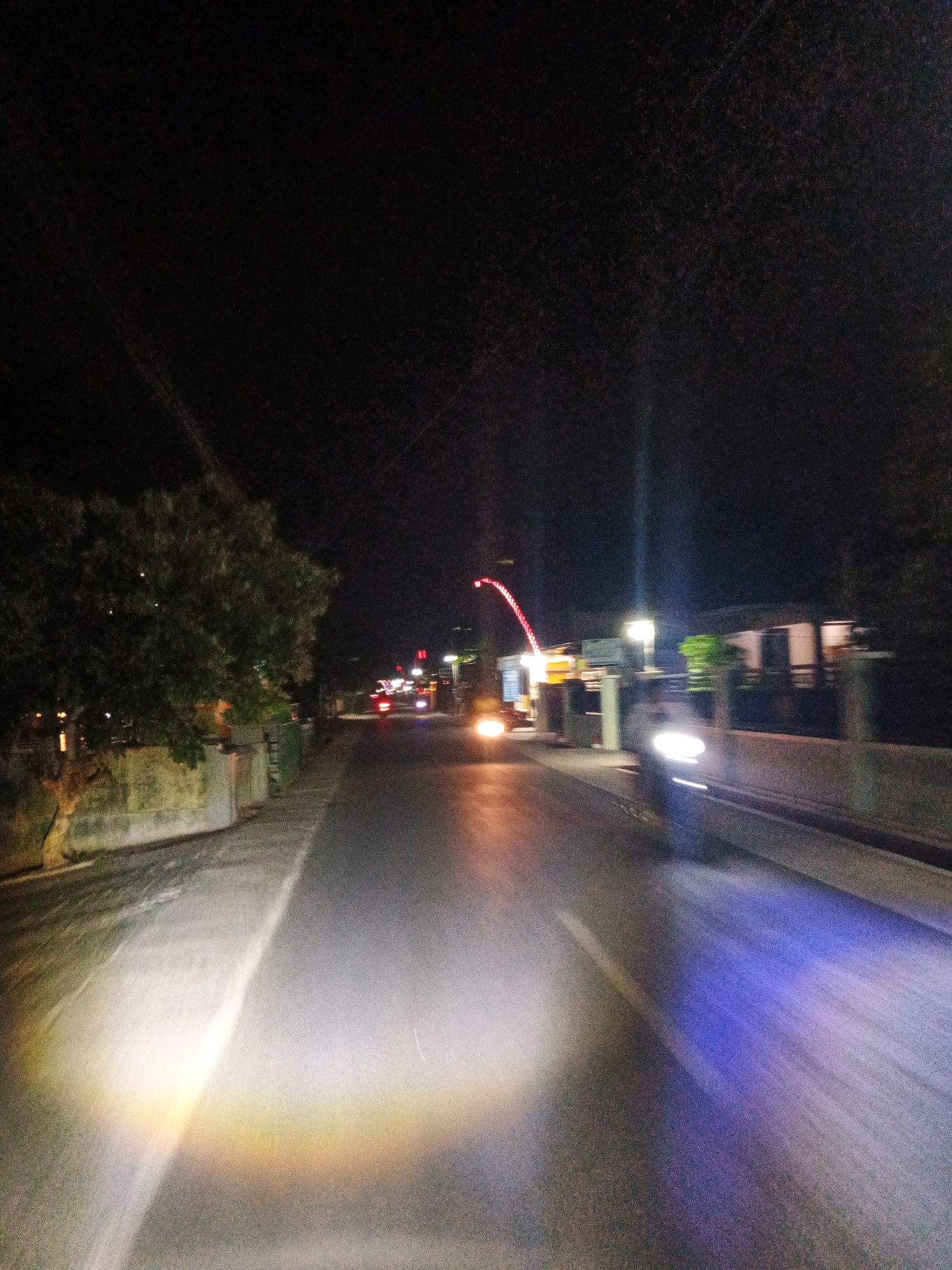 This is a nighttime, slightly motion-blurred image of a street in Sirau, Indonesia. The perspective is from a moving vehicle, likely a motorcycle given the strong headlight beams and the blue light splash on the right side of the road. The road is paved and appears to be a two-lane street. On the left side of the road, there is a low wall with a textured surface, followed by dense foliage from a tree. Further down the street, there are streetlights and buildings visible, with some lights on inside. The right side of the road has a raised barrier or sidewalk. The sky is dark and clear. Traffic lights and vehicle taillights are visible in the distance, indicating ongoing activity on the street. The overall impression is of a typical urban street scene at night, captured while in transit.