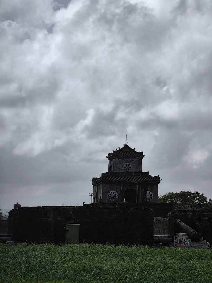 An exterior view depicts a historical fortification in Phú Xuân Ward, Vietnam, under a heavily overcast sky. A two-tiered, dark stone or brick tower, characteristic of the Hue Citadel, dominates the midground, rising above a substantial, dark fortification wall. The tower features a dark tiled roof, multiple arched openings, and circular decorative elements on its facades, capped by a slender central finial. To the tower's immediate left, a light green rectangular panel, possibly a utility box or door, is affixed to the lower wall. To the right, a dark, large-caliber cannon rests on a marked stone base adjacent to the wall, oriented towards the right. A smaller, darker tower or turret is faintly visible in the distant left background atop the extended fortification. The foreground consists of a dense field of vibrant green grass. The sky is largely obscured by extensive, uniform grey clouds, with a minor brighter patch visible in the upper left quadrant. No individuals or discernible activities are present within the scene.