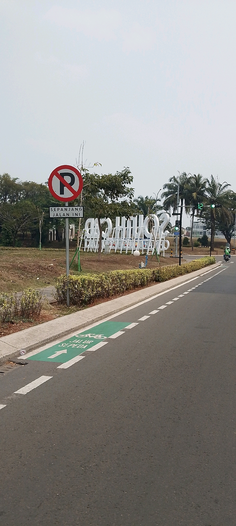 The image depicts a street scene in Gunung Putri, Indonesia, focusing on a dedicated bicycle lane marked in green on the asphalt road. The lane features white directional arrows and the Indonesian text "JALUR SEPEDA" indicating "Bicycle Lane". A circular "No Parking" sign with a red border and a diagonal red line over a black "P" is prominently displayed on a metal pole, accompanied by a smaller rectangular sign below that reads "SEPANJANG JALAN INI", meaning "Along This Road". In the background, large white three-dimensional letters spell out "SOUTH CITY", likely a development or area name, with a row of white benches or seating structures behind them. Palm trees and other foliage are visible, suggesting a tropical or subtropical environment. A traffic light with a green signal and a digital display showing "12" is also present. A person on a scooter, wearing a green helmet, is seen riding away from the camera on the right side of the road. The sky is overcast.