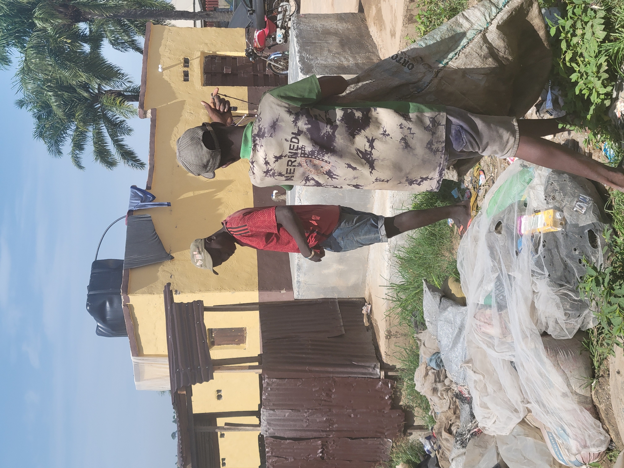 The image depicts a scene in the city of Kwamba, Nigeria, where two young men are engaged in sorting through a pile of discarded materials. In the background, a yellow building with a corrugated metal roof and a water tank is visible. The ground is littered with various types of waste, including plastic bags, fabric scraps, and what appears to be a large sack.
One of the men, wearing a red t-shirt and denim shorts, stands to the left, looking down at the pile. The other man, on the right, wears a camouflage-patterned shirt and grey shorts, and is holding a large, woven sack. His shirt has the word "NERNEDL" printed on the back. The environment appears somewhat neglected, with overgrown grass and debris scattered around. The presence of palm trees suggests a tropical climate. A motorcycle is parked to the right of the building. The overall impression is one of informal waste management or scavenging.