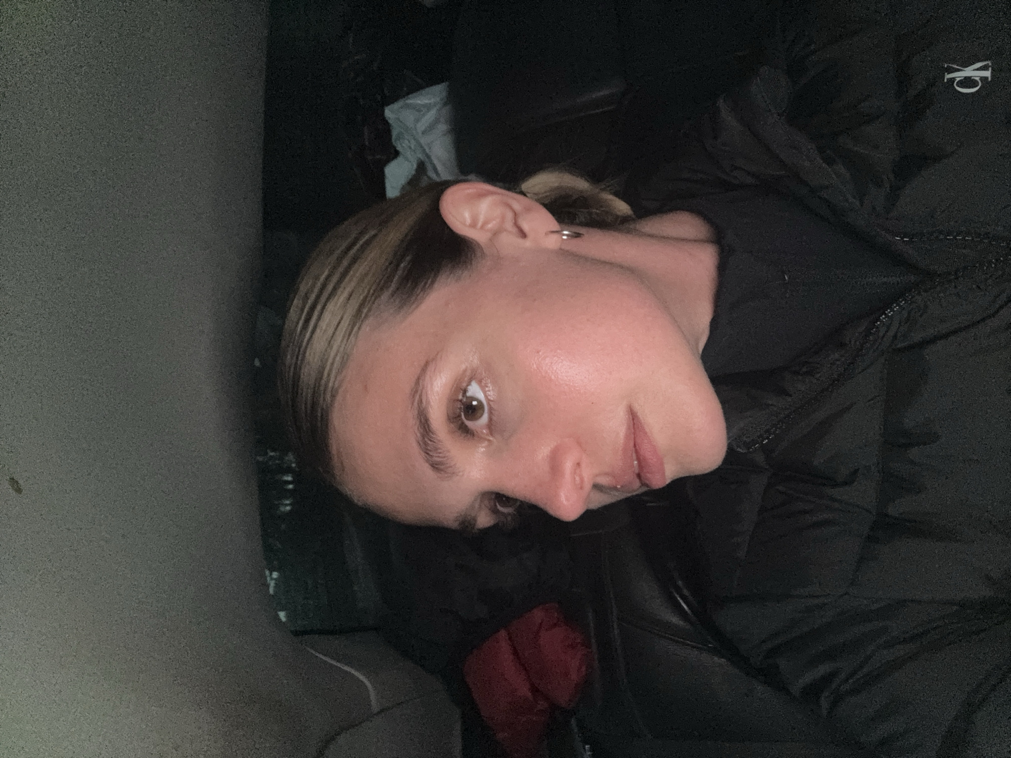 A young woman is depicted in a portrait-style image, rotated 90 degrees clockwise, captured in low light. She has light brown hair, pulled back from her face, and brown eyes. Her expression is neutral, and her lips are slightly parted. A small, silver hoop earring is visible in her left ear.
She is wearing a black puffer jacket, which features a white "CK" logo, indicating Calvin Klein, on the upper left chest area. The background is predominantly dark and out of focus, suggesting an indoor or low-light setting, possibly within a vehicle or a dimly lit room. A textured, grey-brown surface is visible on the left side, and a hint of a red object, possibly a piece of clothing, can be seen near her body in the lower central part of the frame. The lighting on her face suggests a direct, possibly artificial light source, creating a slight sheen on her skin. She appears to be posing for the camera.