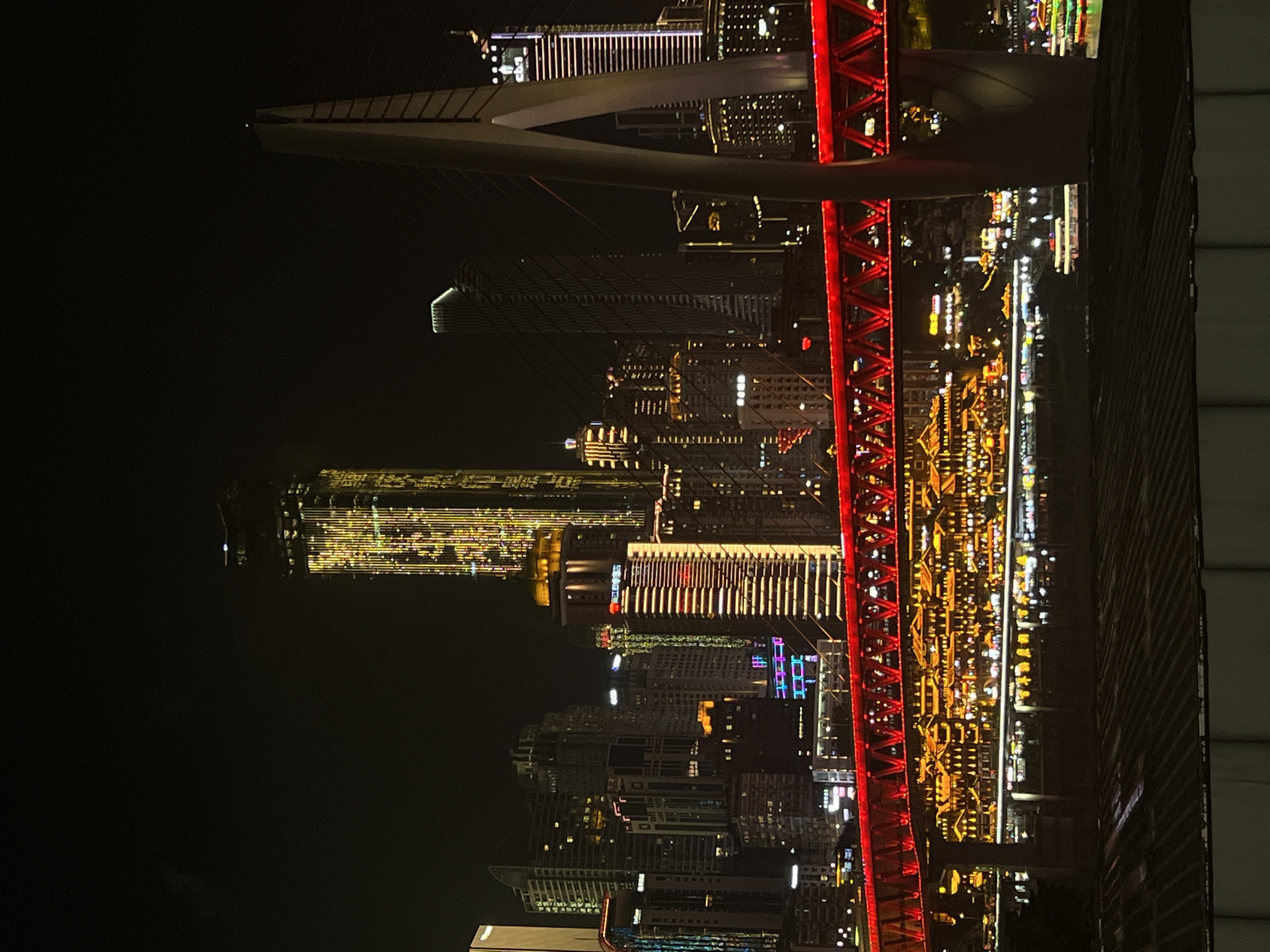 The image shows a nighttime cityscape with a prominent red bridge in the foreground. The bridge is illuminated with a vibrant red light, highlighting its intricate metal structure. Beyond the bridge, a dense collection of skyscrapers pierces the dark sky, their windows aglow with various colors of light, predominantly yellow and white. Some buildings are lit up in patterns, suggesting architectural illumination.
The scene depicts a bustling urban environment at night. The time of day is clearly nighttime, as indicated by the darkness of the sky and the artificial illumination of the city. There are no visible people or specific activities taking place, suggesting the image is a general view of the city. The setting appears to be a waterfront area, as suggested by the reflections of lights that could be on water, and the arrangement of buildings. The overall mood conveyed is one of urban vitality and modern architectural grandeur.
There is some visible text on one of the buildings in the middle distance, appearing as red characters, possibly a business name or advertisement, but it is not legible. The location context provided is Jiangbeicheng Subdistrict, China, which aligns with the appearance of a major Chinese urban center. The image is oriented vertically, with the bridge and city appearing upside down relative to a typical horizontal view.