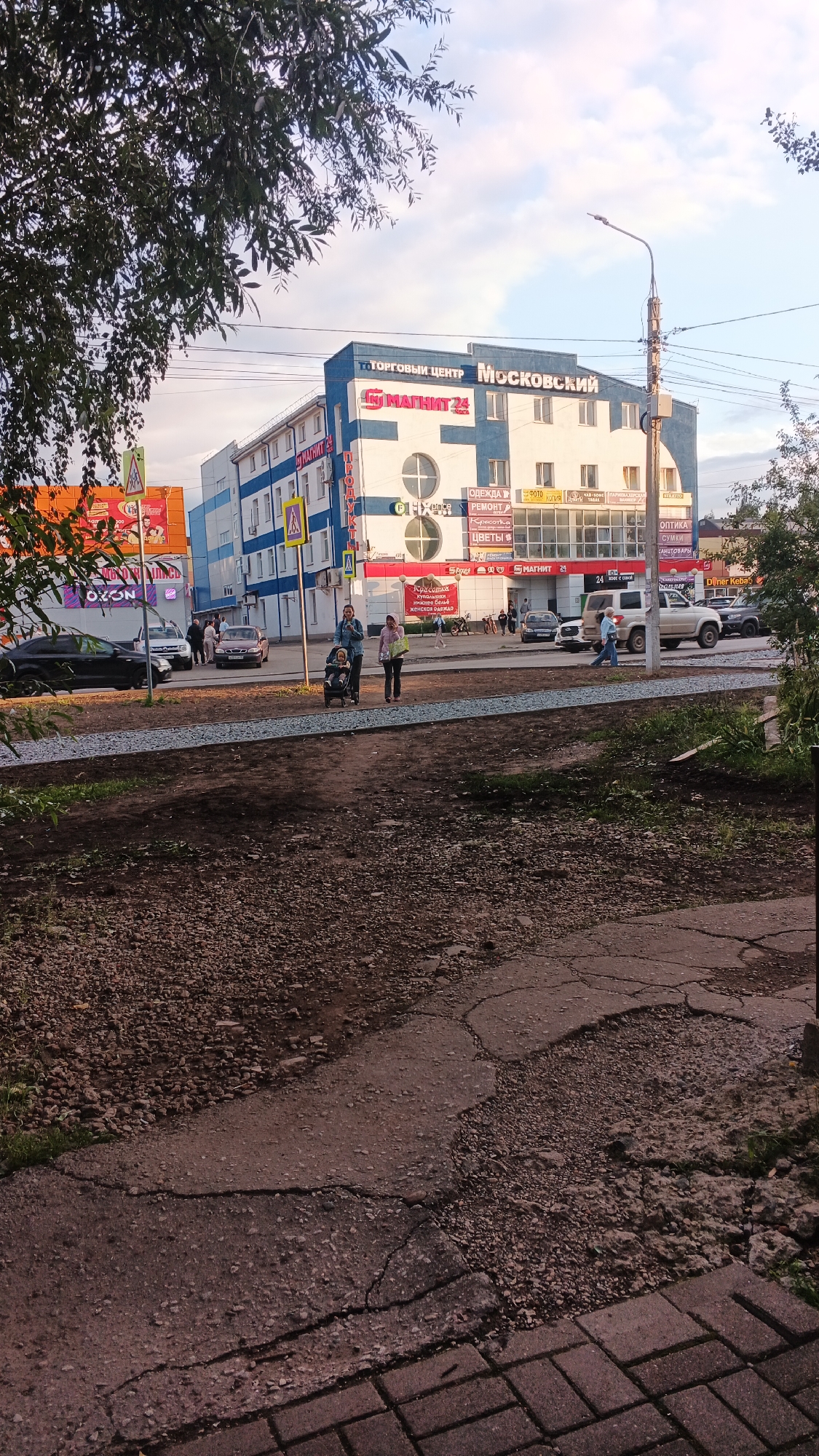 The image shows a street scene in Izhevsk, Russia, on a bright, cloudy day. In the background, a multi-story building with blue and white facade stands prominently, featuring a sign for "ТОРГОВЫЙ ЦЕНТР Московский" (Shopping Center Moskovsky). Below this, a large red and white sign displays "МАГНИТ 24" (Magnit 24), a Russian supermarket chain. Various shop signs are visible on the ground floor, including "ОДЕЖДА" (Clothing), "РЕМОНТ" (Repair), "КРАСОТКА" (Beauty), "ЦВЕТЫ" (Flowers), and "ОПТИКА" (Optics).
In the middle ground, a woman with a baby in a stroller and another woman are walking on a paved area. Several cars are parked along the street and on the road, including a white SUV, a dark sedan, and a black sedan. A traffic light and a street lamp are also visible.
The foreground depicts a rough, unpaved area with gravel and some patchy vegetation, transitioning into cracked asphalt and then brick paving. The overall environment suggests an urban setting with elements of commercial activity and public infrastructure.