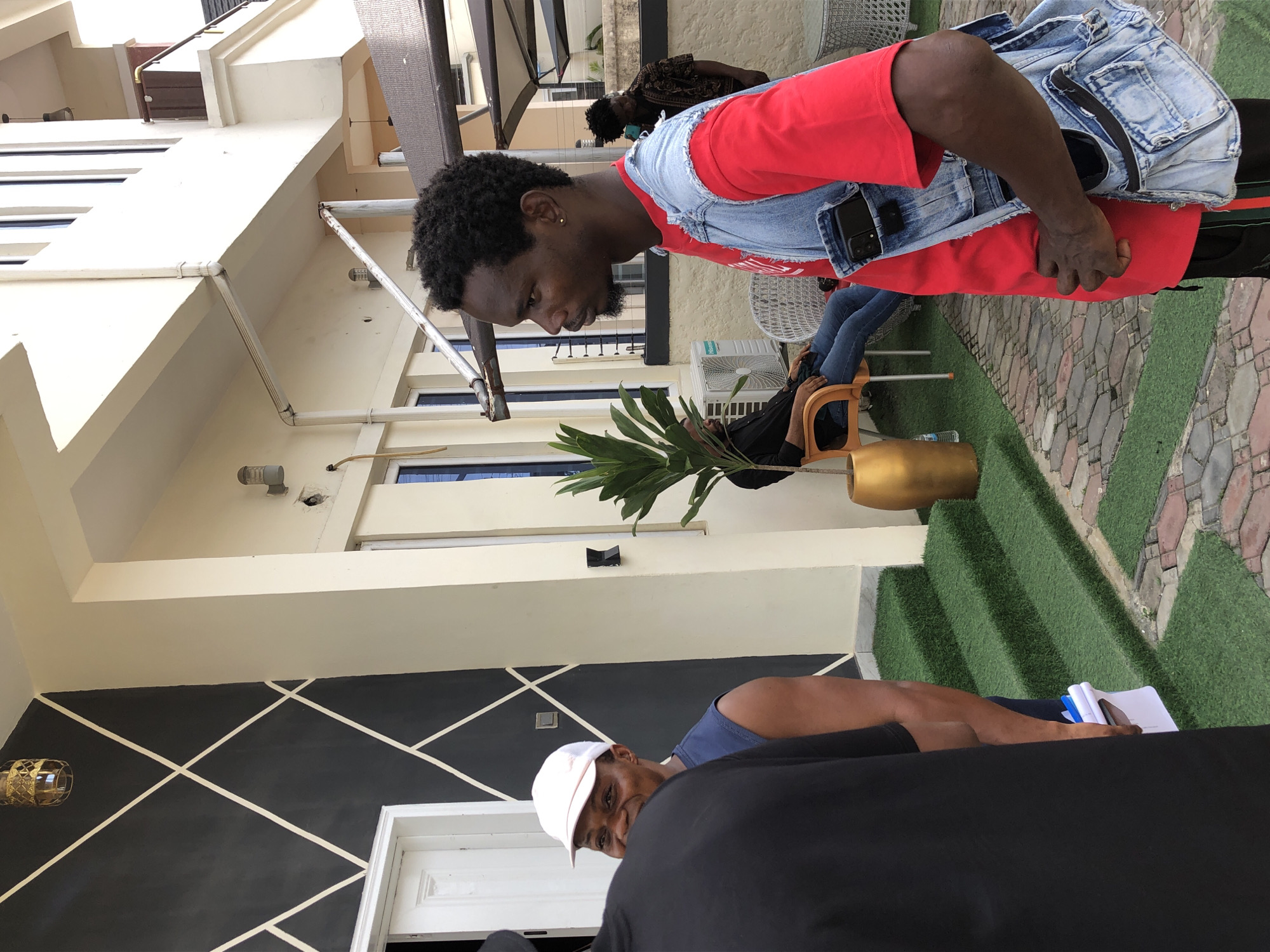 A candid, daylight photograph captures several men in an outdoor setting, likely a modern residential or commercial courtyard. The image is oriented vertically (originally rotated 90 degrees clockwise), showing a multi-story building with light-colored walls and a distinctive black accent wall featuring white diagonal geometric patterns. Artificial green grass steps lead up to the building, alongside a paved area of interlocking pavers.
In the mid-ground, a dark-skinned man with short, curly hair and a beard is prominent. He wears a long-sleeved red shirt under a light blue denim vest, with a black phone case visible in his vest pocket. He is standing, leaning forward and looking downwards to his left. His expression is focused.
To the lower left, partially obscured by another person's black clothing, another dark-skinned man is visible. He wears a white baseball cap and a blue tank top, looking directly at the camera with a slight smile. Next to him, a small white notebook and pen are visible.
In the background, behind the primary subject, another man sits casually on a light brown chair, wearing a black shirt and jeans. An outdoor fan unit is visible near him, and a large plant in a golden pot stands nearby. Further back, another person in a patterned shirt is partially visible.
The scene suggests a relaxed social gathering in bright, sunny weather. There is no visible text.