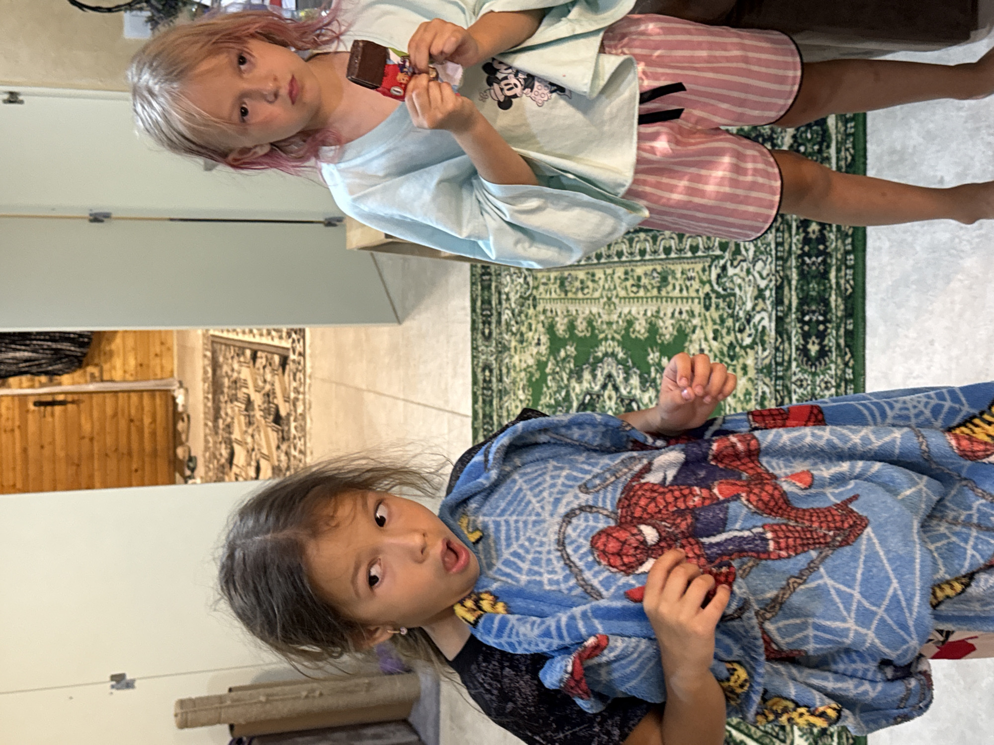 Two young girls are standing in a room with wooden walls and a patterned rug. The girl on the left is wearing a black t-shirt and is holding a blue towel with a Spider-Man print. She has her mouth open and is looking to the left. The girl on the right is wearing a light blue shirt with Mickey Mouse printed on it and pink striped shorts. She is holding a chocolate bar and looking forward with a serious expression. The environment appears to be a home.