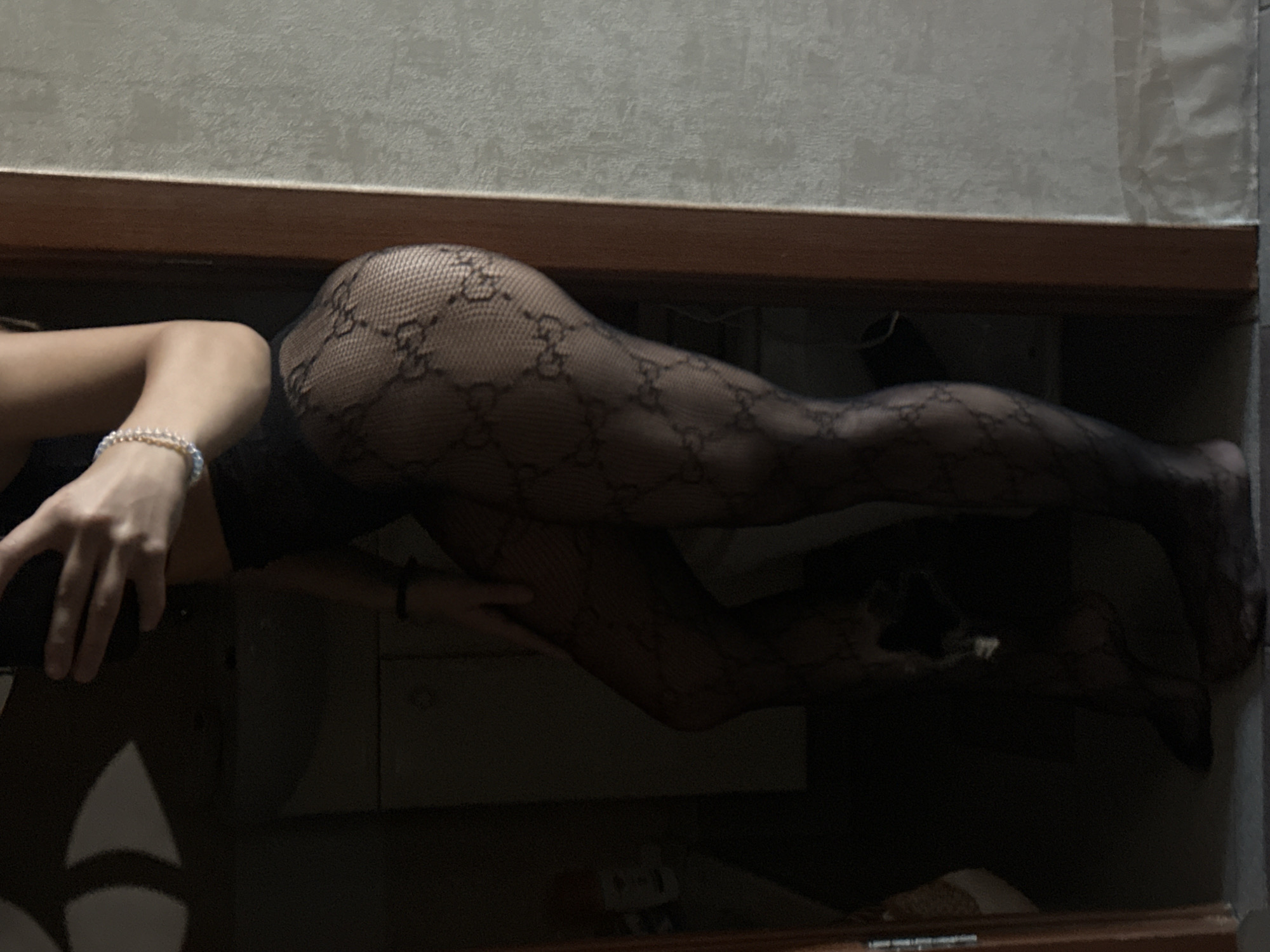 The image displays a person's lower body and hand, positioned horizontally across the frame in an indoor setting located in Irkutsk, Russia. The individual is wearing black fishnet stockings featuring a repeating pattern of dark, interlocking, G-like symbols. A light-colored, possibly beaded, bracelet adorns the person's wrist, and their hand rests on a dark surface.
The upper portion of the image shows a light-colored wall with a visible wooden trim or shelf below it. The main subject, including the legs and hand, appears to be resting or posing on a dark, reflective surface, possibly a polished table or a large mirror. This reflective surface creates a clear, dark reflection of the person's lower body underneath them, extending downwards. The lighting in the scene is dim, suggesting it is evening or night. No specific activities beyond posing are discernible, and no visible text is present in the image.