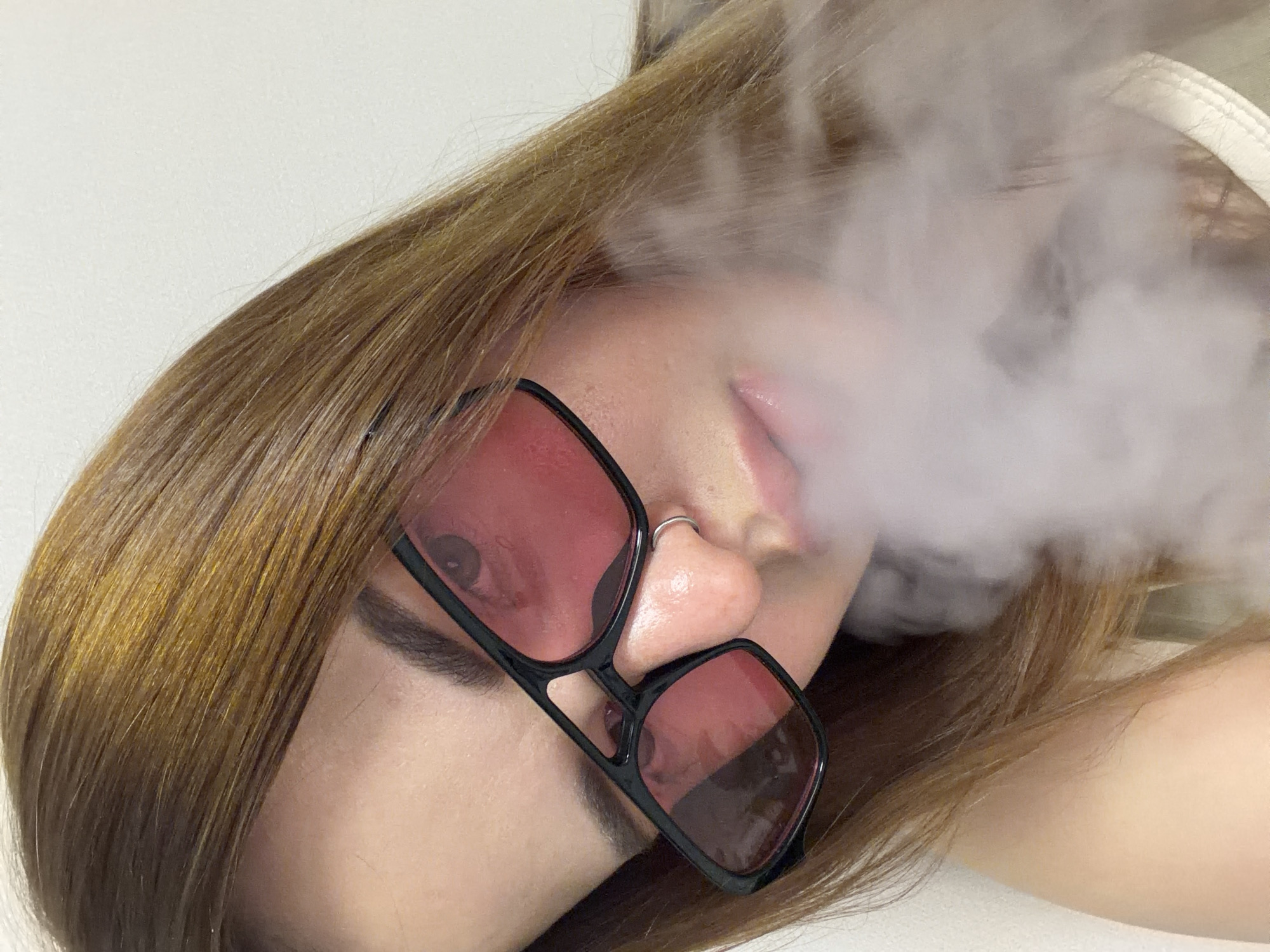 A close-up, angled shot features a person with light brown hair, wearing square-framed sunglasses with reddish-pink tinted lenses. The individual is positioned diagonally, with their head tilted. A significant plume of white smoke or vapor billows around the lower right side of their face, partially obscuring their mouth and cheek.
The person has a silver nose ring on the right side of their nose. Their hair, which appears long and straight, frames the left side of the image and extends towards the bottom. The skin tone is fair. The sunglasses have thick black frames, adding a distinctive element to their look.
The background is a plain, light color, suggesting a minimalist or indoor setting. The lighting is soft and even, highlighting the details of the person's features and the texture of the smoke. The person's expression is largely hidden by the sunglasses and smoke, but their lips are slightly parted. There is no visible text in the image.