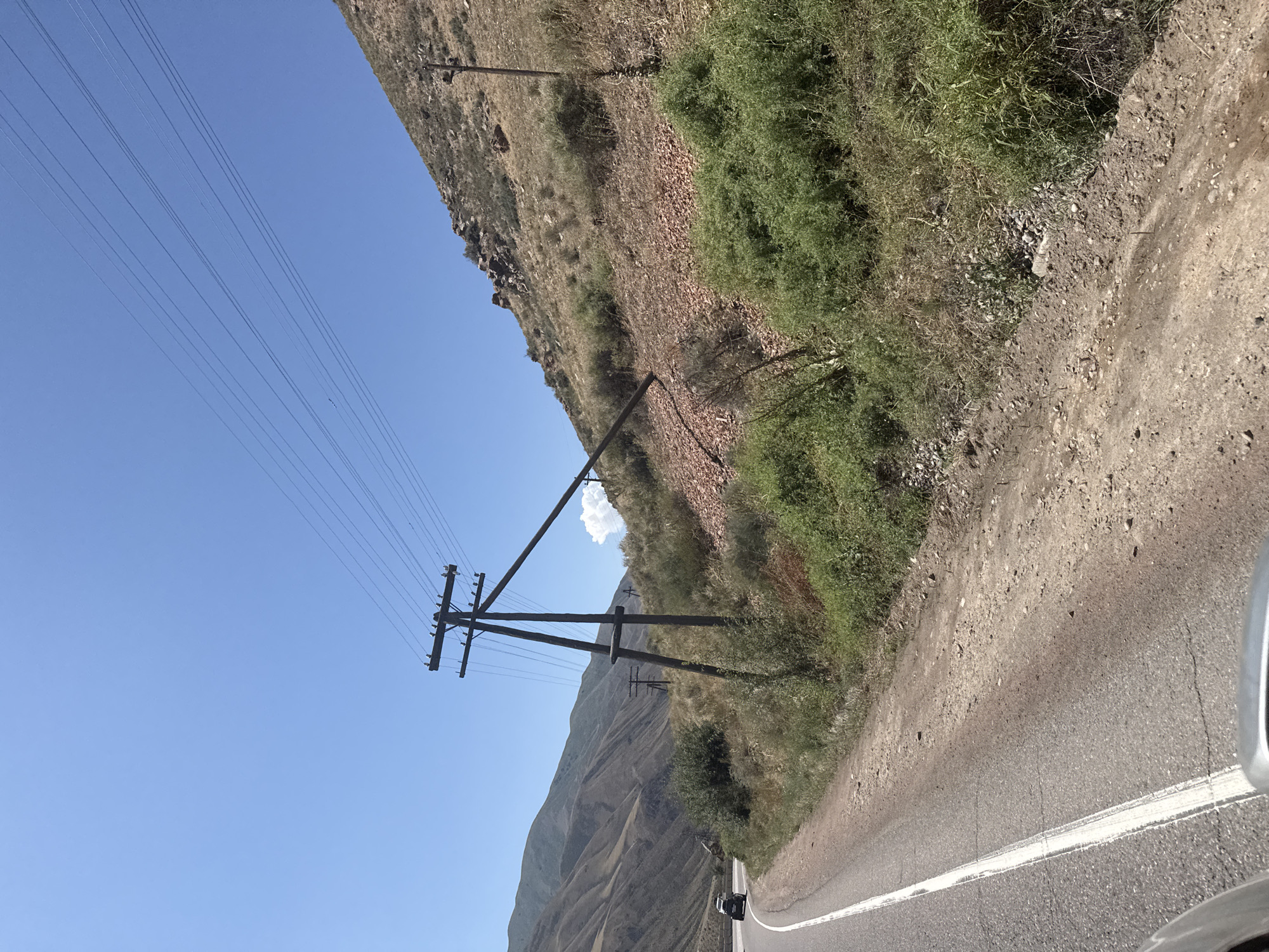 The image shows a roadside scene in the city of Cholok, Kyrgyzstan. A paved road curves through a hilly, arid landscape under a clear blue sky. On the right side of the road, a large wooden utility pole leans precariously, with its crossbar angled downwards, supporting multiple power lines that stretch across the frame. Another utility pole stands further up the slope. The terrain is covered with sparse dry grasses, shrubs, and loose rocks. In the distance, the rolling hills of the surrounding mountains are visible. A dark-colored vehicle is driving on the road in the left portion of the image. The overall impression is one of a rural or semi-rural setting, possibly undergoing or having undergone some environmental stress indicated by the dry vegetation and the damaged utility pole.