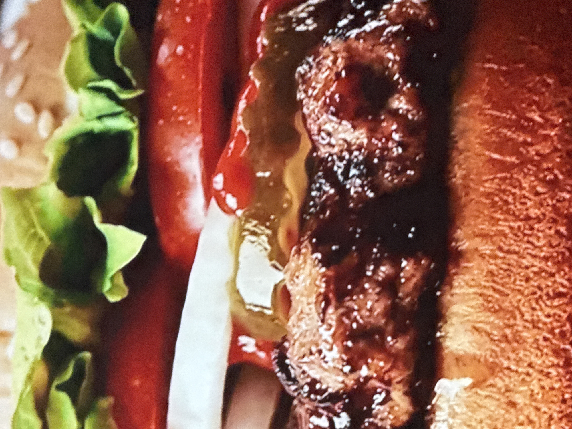 A close-up image shows a cross-section of a hamburger. From left to right: a sesame seed bun, green lettuce, a red tomato slice, a white onion slice, a yellow sauce/pickle, a cooked meat patty with a browned exterior, and the top bun. The focus is on the details of the ingredients and their textures. The location in Mytishchi, Russia is not discernible from this image.