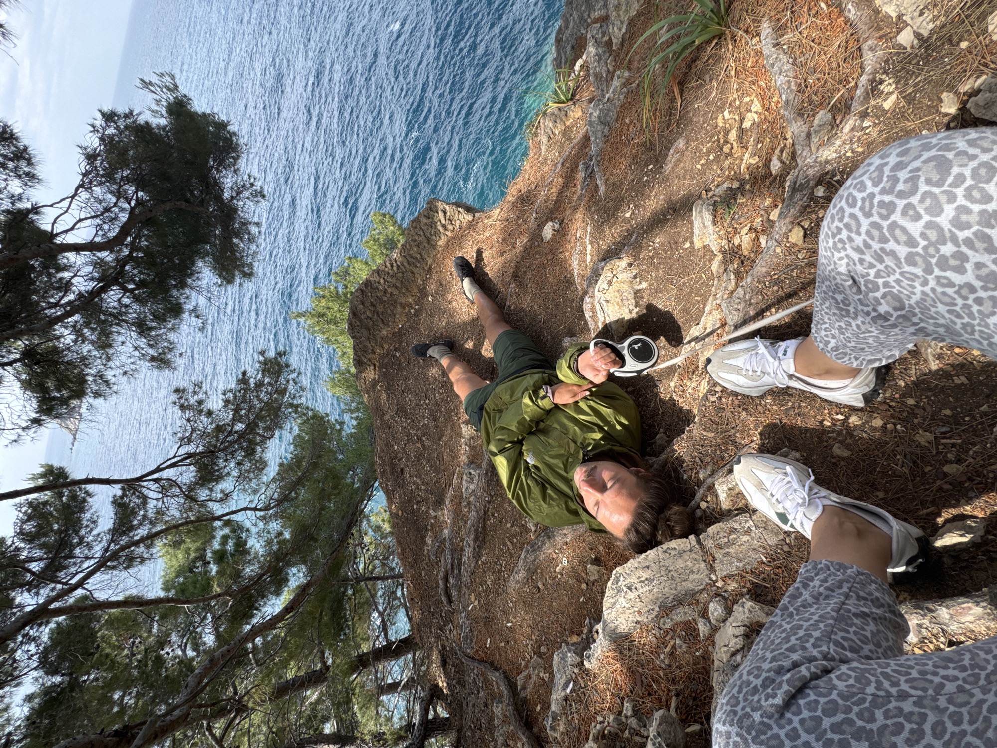 A medium, outdoor shot depicts a man lying on a cliff face near the city of Petrovac na Moru, Montenegro. The cliff edge is to the left, overlooking the Adriatic Sea. The man is positioned in the middle of the frame, wearing a green jacket and dark shorts. He holds a retractable dog leash. The sea is visible in the background, along with trees and vegetation on the cliff. In the foreground, the lower half of another person is visible, wearing spotted leggings and white sneakers. The ground is covered in dirt, rocks, and pine needles. The photo is taken from an elevated perspective, possibly by the person in the foreground. The photo is rotated slightly clockwise.