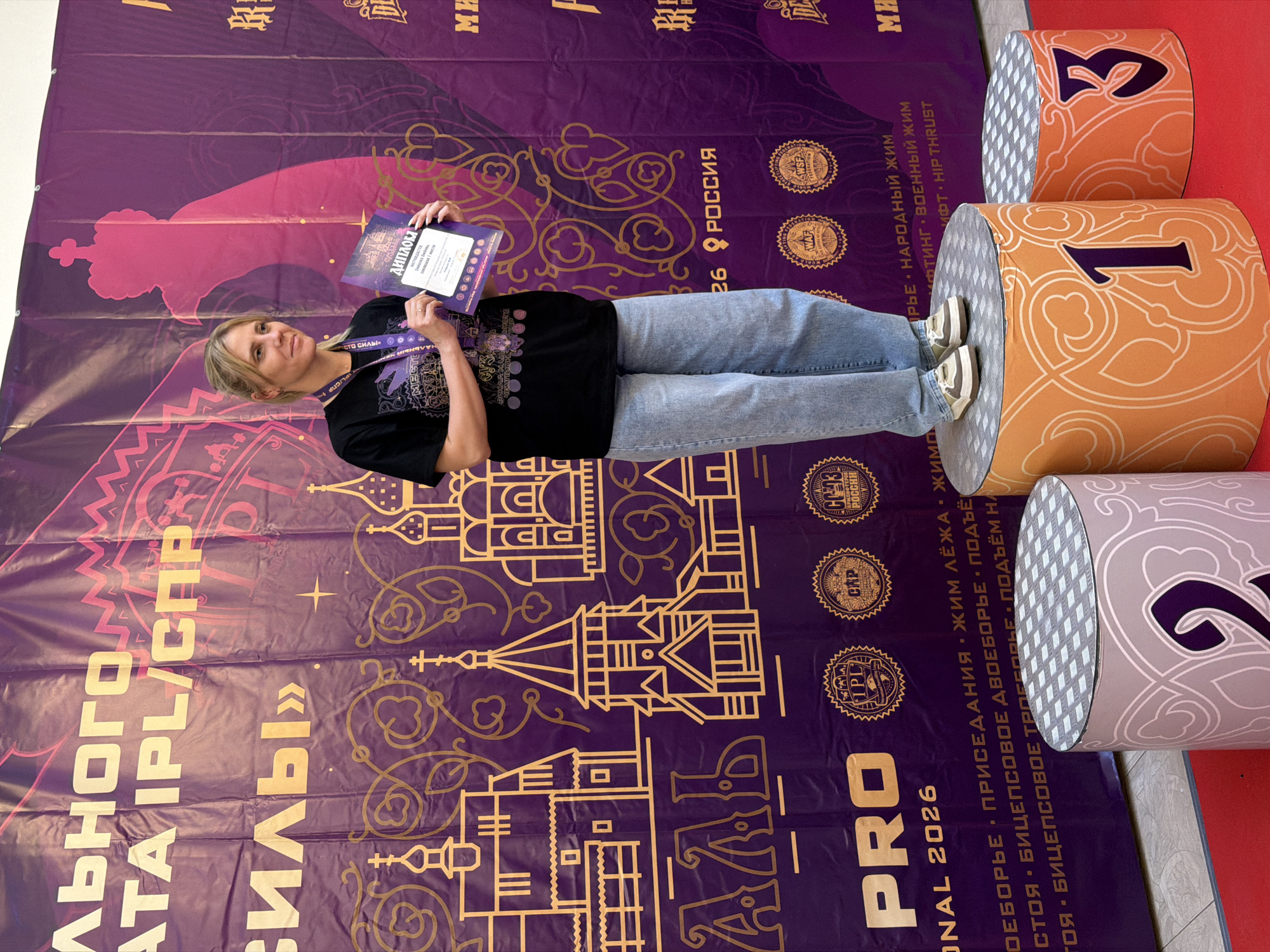 A blonde woman stands on a podium, holding a certificate. She is wearing a black t-shirt and blue jeans. Behind her is a large purple banner with text and illustrations of buildings. The banner reads "ЛЬНОГО АТА IPL/СНР СИЛЫ" and features decorative patterns and images of Russian Orthodox churches. There are also logos for various organizations on the banner, including "IPL", "WAP", "WSF FEDERATION", and "GC-93 РОССИЯ". The woman is positioned in front of a silhouette of a crown. The podiums are cylindrical and decorated with orange and purple patterns, with the numbers "1" and "3" visible on two of them. The scene appears to be a competition or award ceremony.