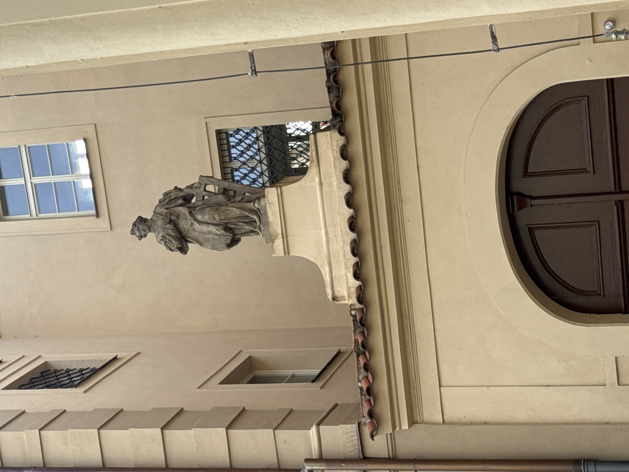 The image shows a statue of a man in historical attire, standing on a pedestal atop a building in Prague, Czechia. The statue is made of stone and depicts the man holding a scroll or book. The building has a light beige facade with red-tiled roofing and arched doorways. There are several windows visible, some with intricate metal grilles. The overall scene suggests an architectural detail from an older part of the city, likely a historical site or religious building.