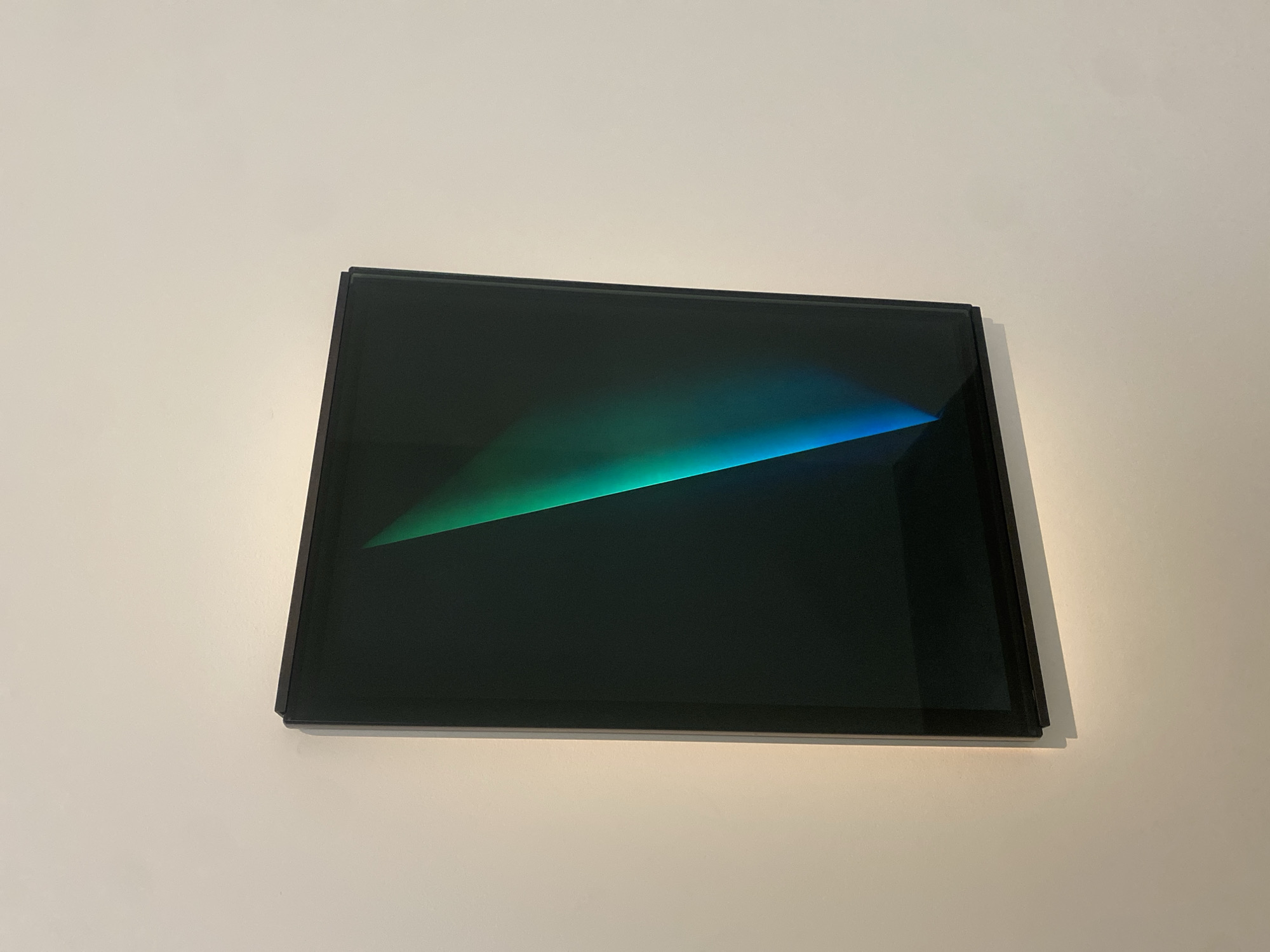 A black rectangular artwork hangs on a stark white wall. The artwork features a gradient of colors, transitioning from a vibrant green on the left to a deep blue on the right, all against a dark background. The artwork appears to be slightly angled away from the wall, casting a subtle shadow. The scene does not reveal any identifying information about the location in Paris, France. The wall appears to be clean and unmarked.